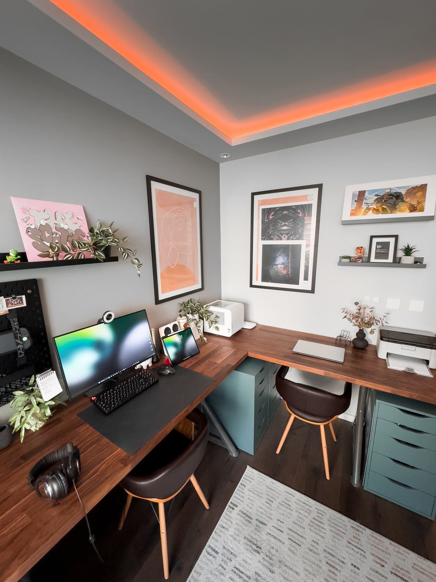 A modern home office setup with a Dell monitor, Razer Kiyo webcam, Logitech G Pro keyboard and mouse, an iPad Pro 12.9 (3rd Gen), Logitech G Pro X headphones, and a printer, all on a wooden IKEA KARLBY desk with IKEA ALEX drawers, decorated with plants and framed artwork on the walls