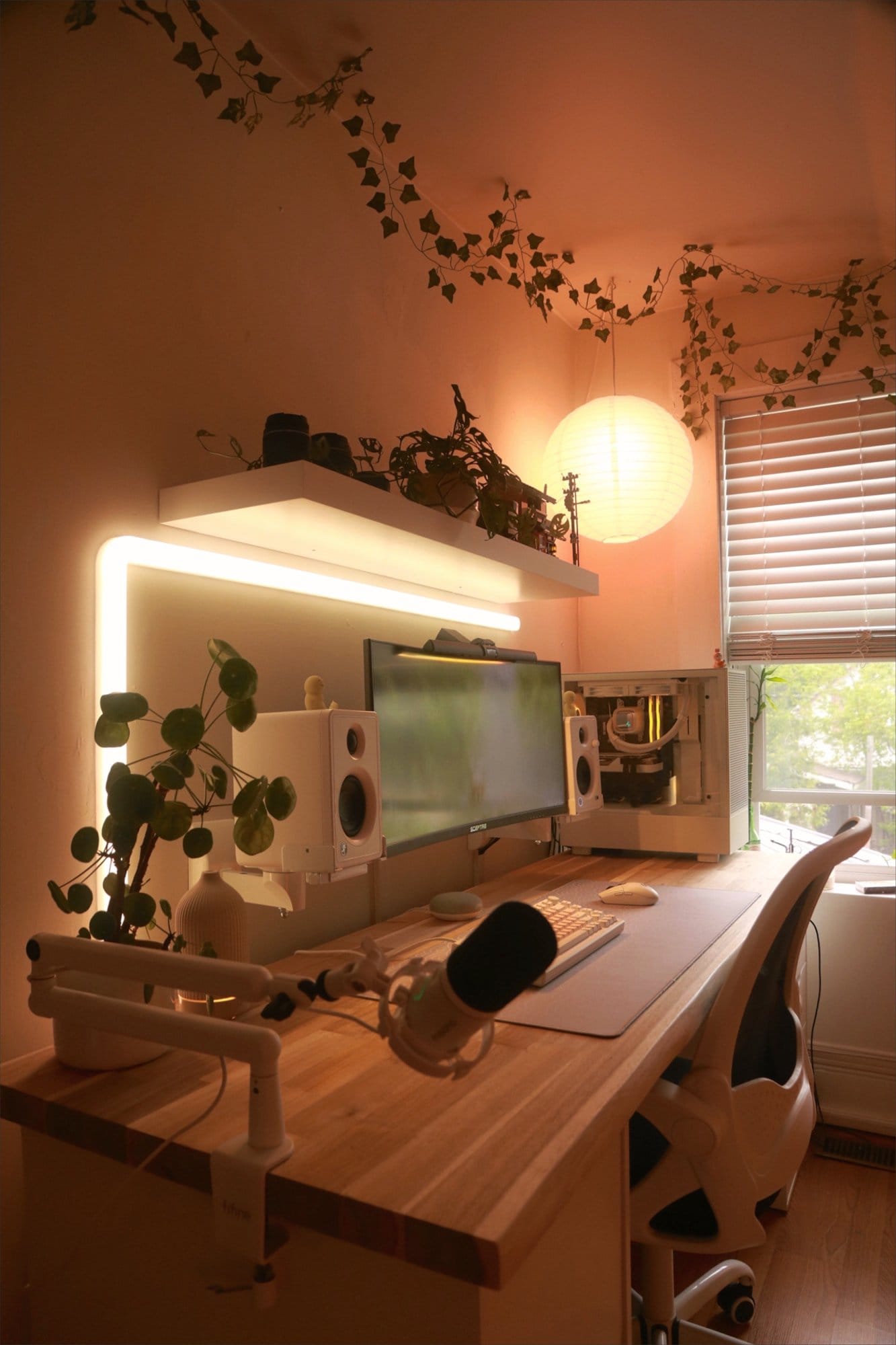A cosy setup with a Sceptre 30″ Ultrawide monitor, Mackie CX3 speakers, and an AKKO MOD 007 PC keyboard on a wooden desk, illuminated by soft ambient lighting and adorned with plants