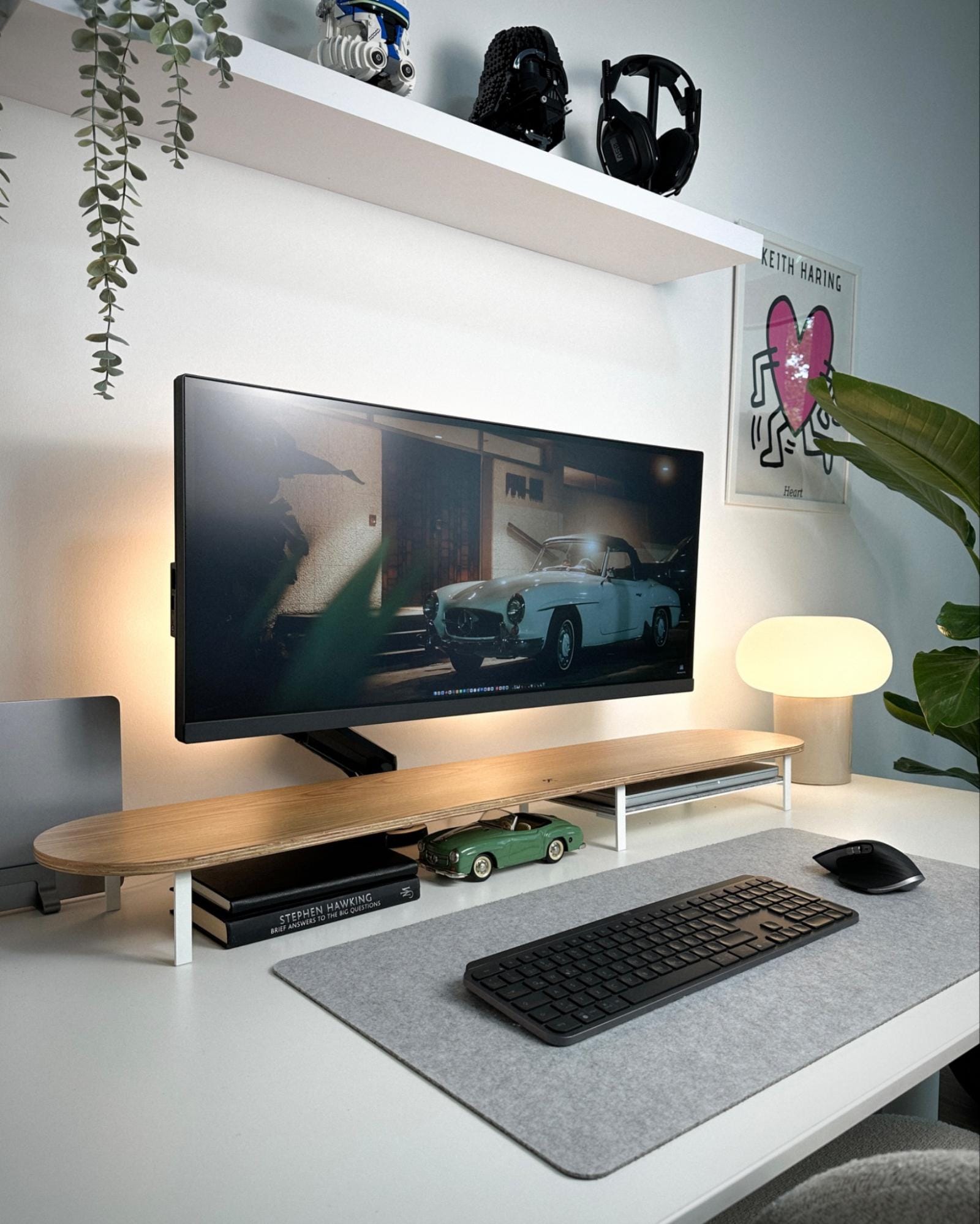 A modern desk setup featuring an ultrawide BenQ monitor, a Logitech MX Master 3S mouse and keyboard on a felt mat, a Woodcessories desk shelf, LEGO Star Wars Darth Vader and Captain Rex helmets on a shelf above, and an Astro A50 headset, with a Keith Haring ”Heart“ poster on the wall and a modern lamp illuminating the corner