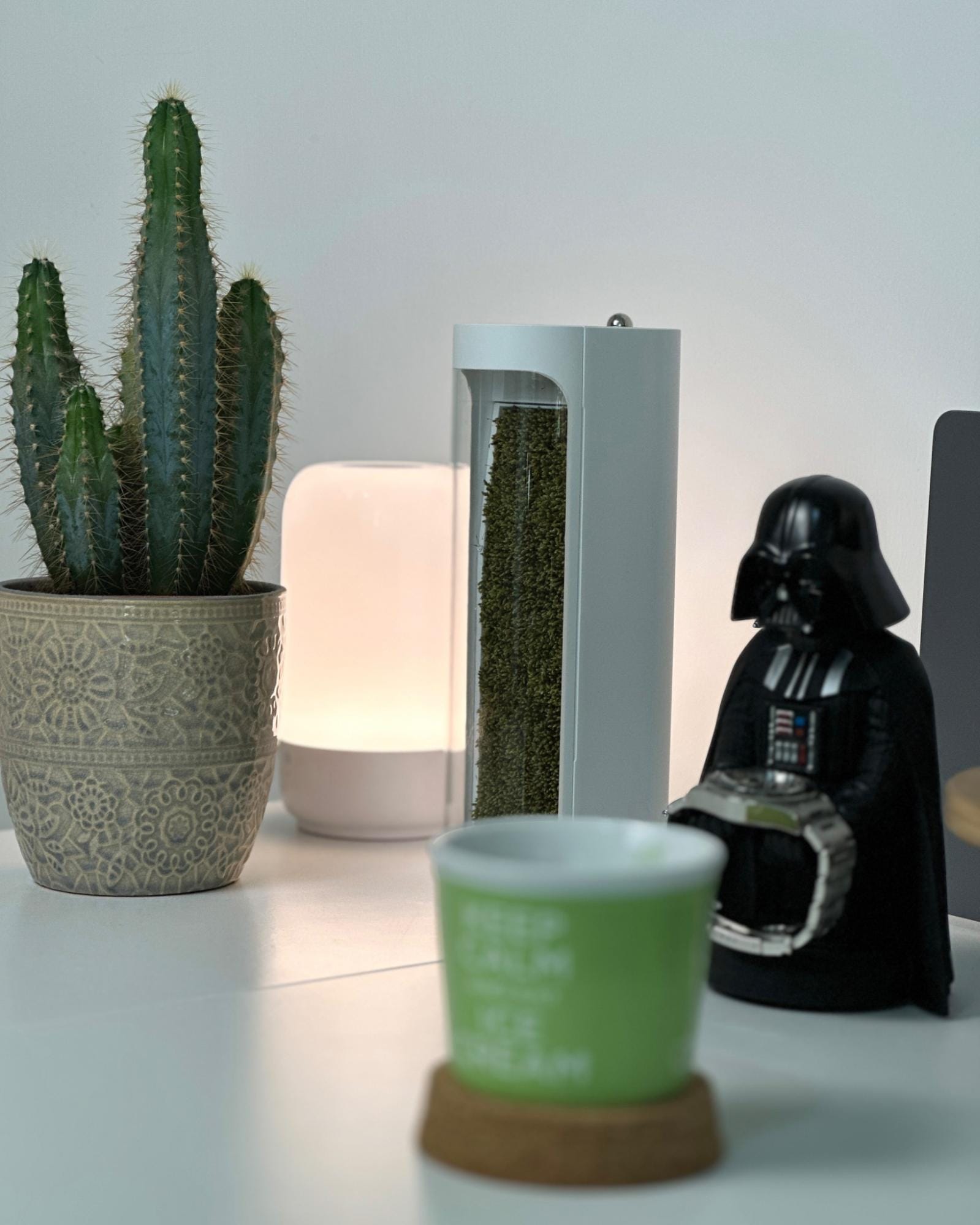 A close-up of a desk setup featuring a cactus in a decorative pot, a modern lamp, a mosslab Moss Air purifier, a Star Wars: Darth Vader Gaming Controller Holder, and a green mug with the text ”KEEP CALM AND EAT ICE CREAM”