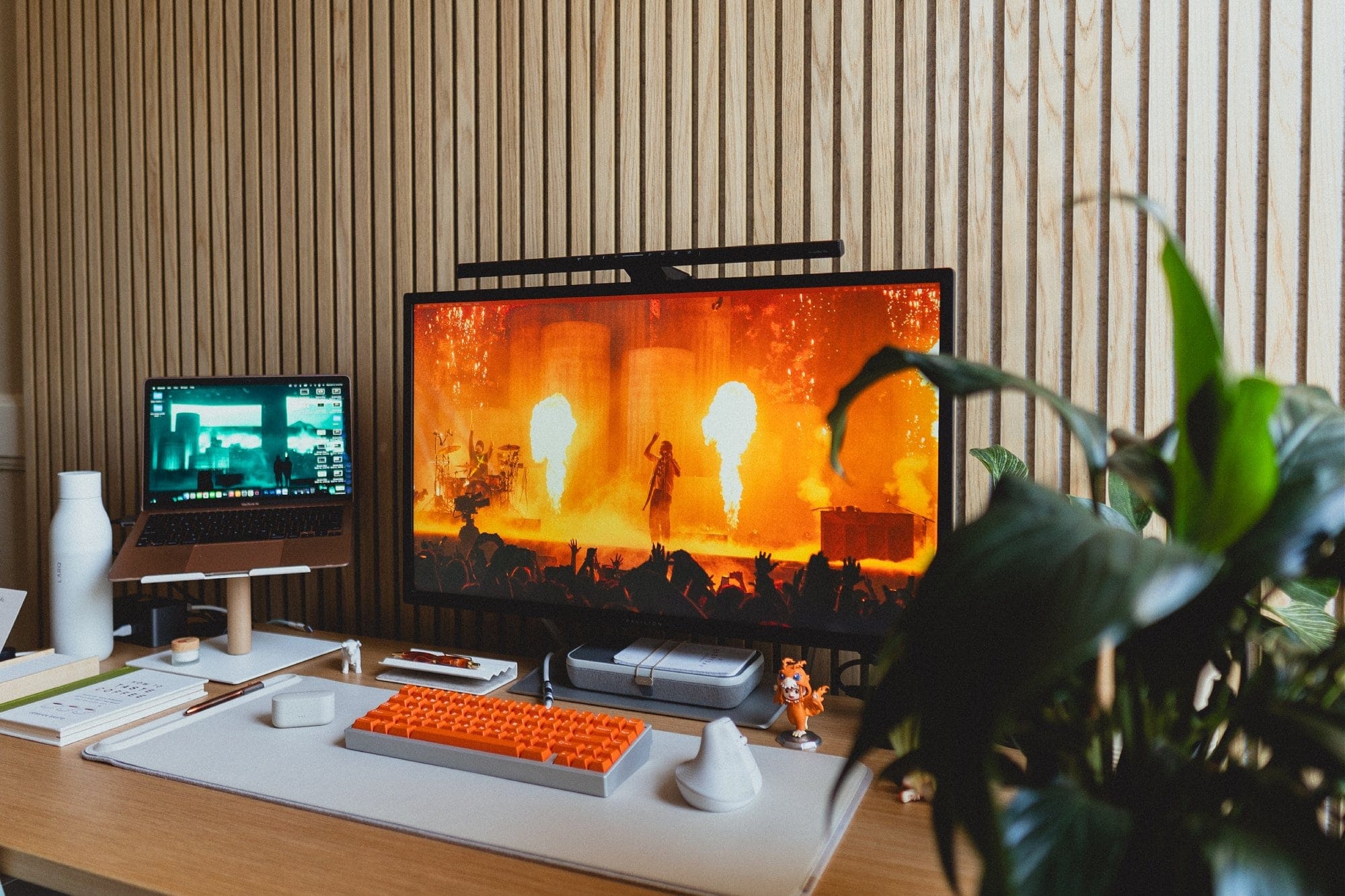 A workspace setup with a M1 MacBook Air on an Ugmonk stand, an HP Pavilion monitor showing a fiery concert scene, a KMG B65 keyboard with bright orange DCX Topaz keycaps, a Logitech Lift mouse, and lush plants in the foreground