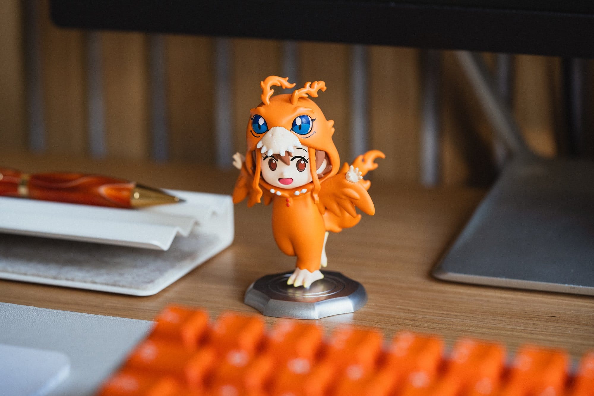 A close-up of a Sora Digimon anime-style figurine on a desk, positioned near a pen holder and partially in front of an orange keyboard