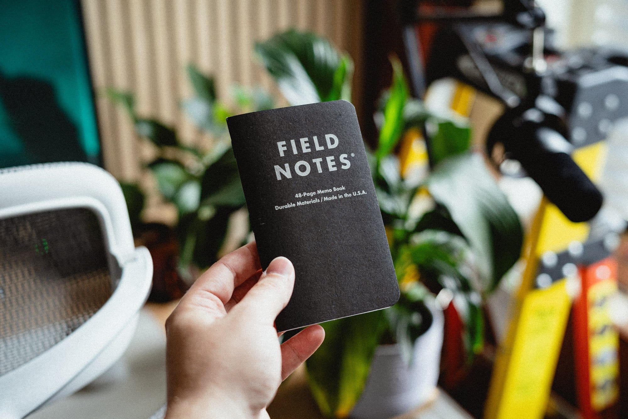 A hand holding a black Field Notes memo book with “48-Page Memo Book, Durable Materials, Made in the U.S.A.“ written on the cover, against a background of plants and desk equipment
