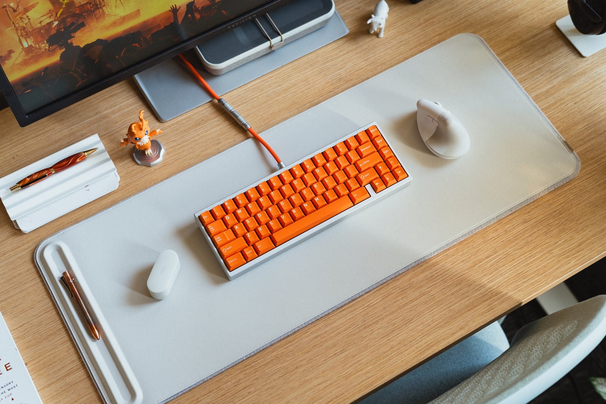 A desk setup with an orange KMG B65 keyboard on a white Orbitkey Slim desk mat, accompanied by a Logitech Lift mouse, a Sora Digimon figurine, and a red and gold pen in a holder