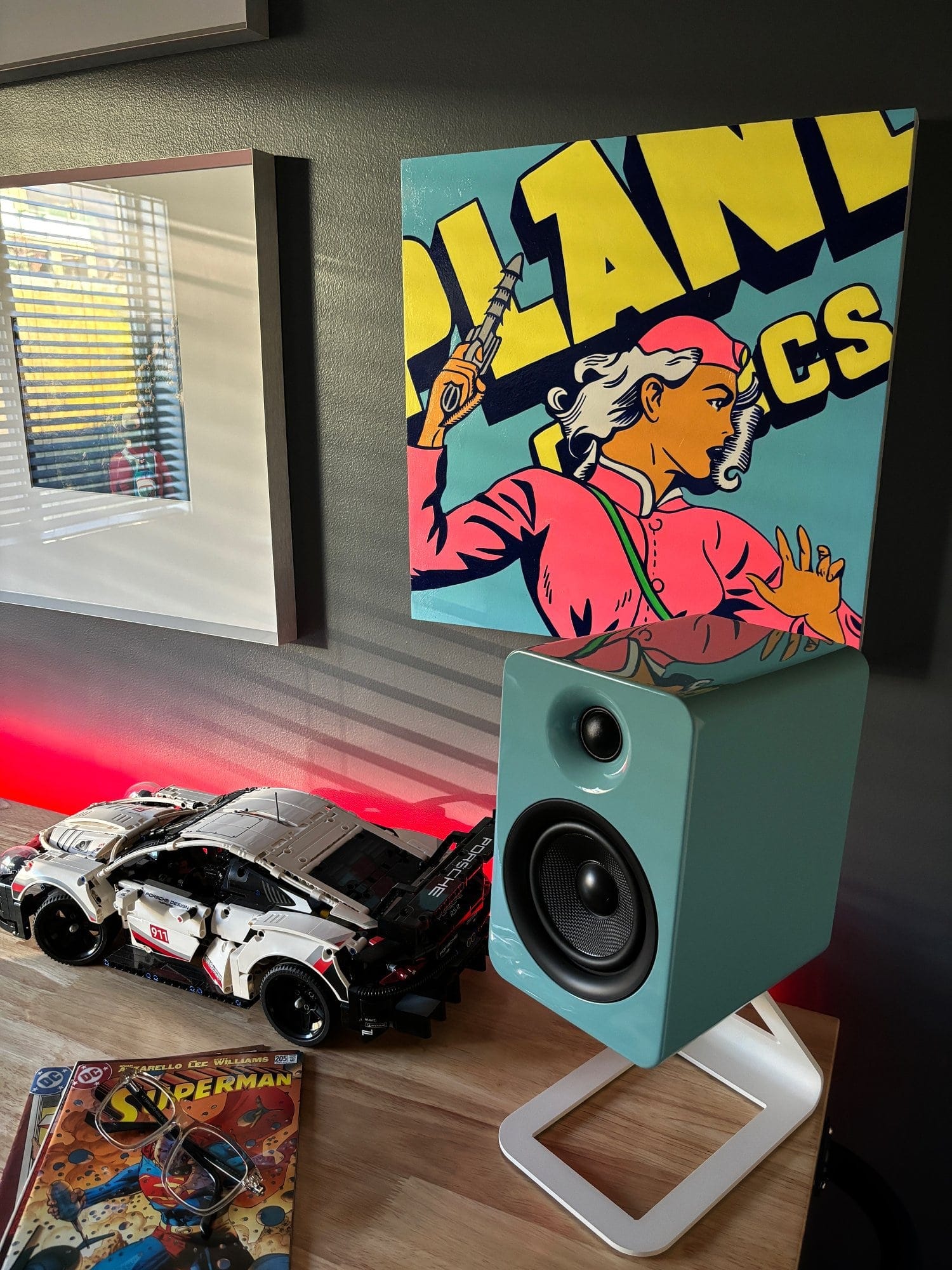A Kanto YU4 speaker in gloss teal on a white stand, a LEGO Porsche model, and vintage comic books on a wooden workbench with red LED lighting