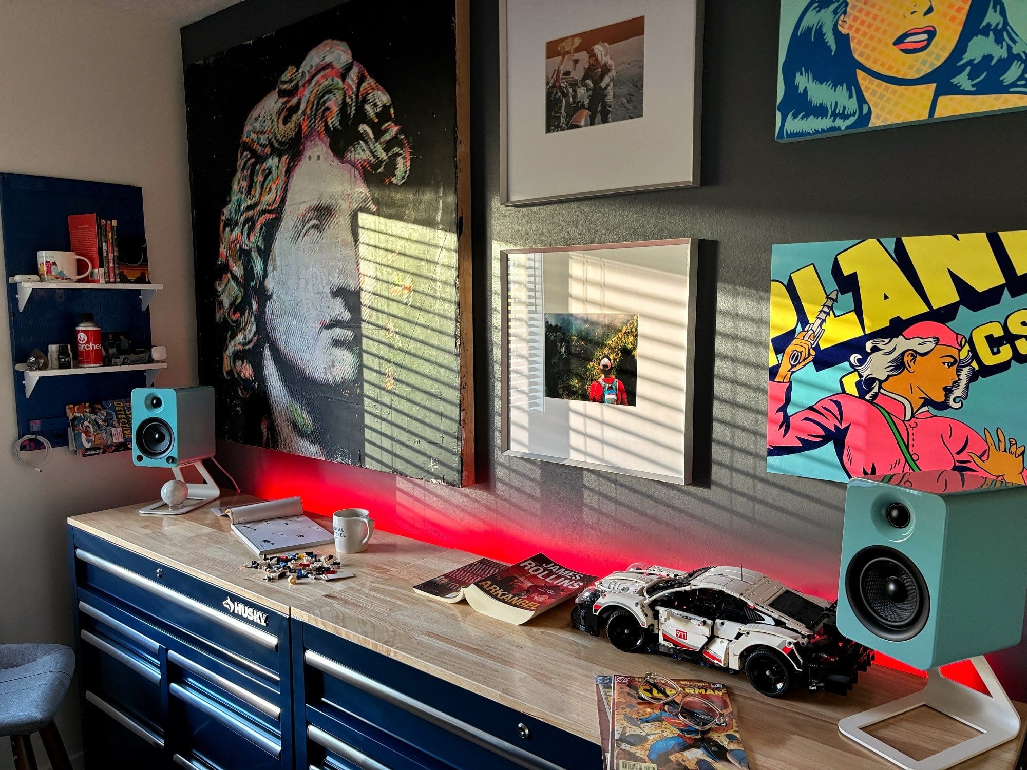 A workspace with a Husky workbench, Kanto YU4 speakers in gloss teal, a LEGO Porsche model, and comic book art on the walls, lit by red LED lighting