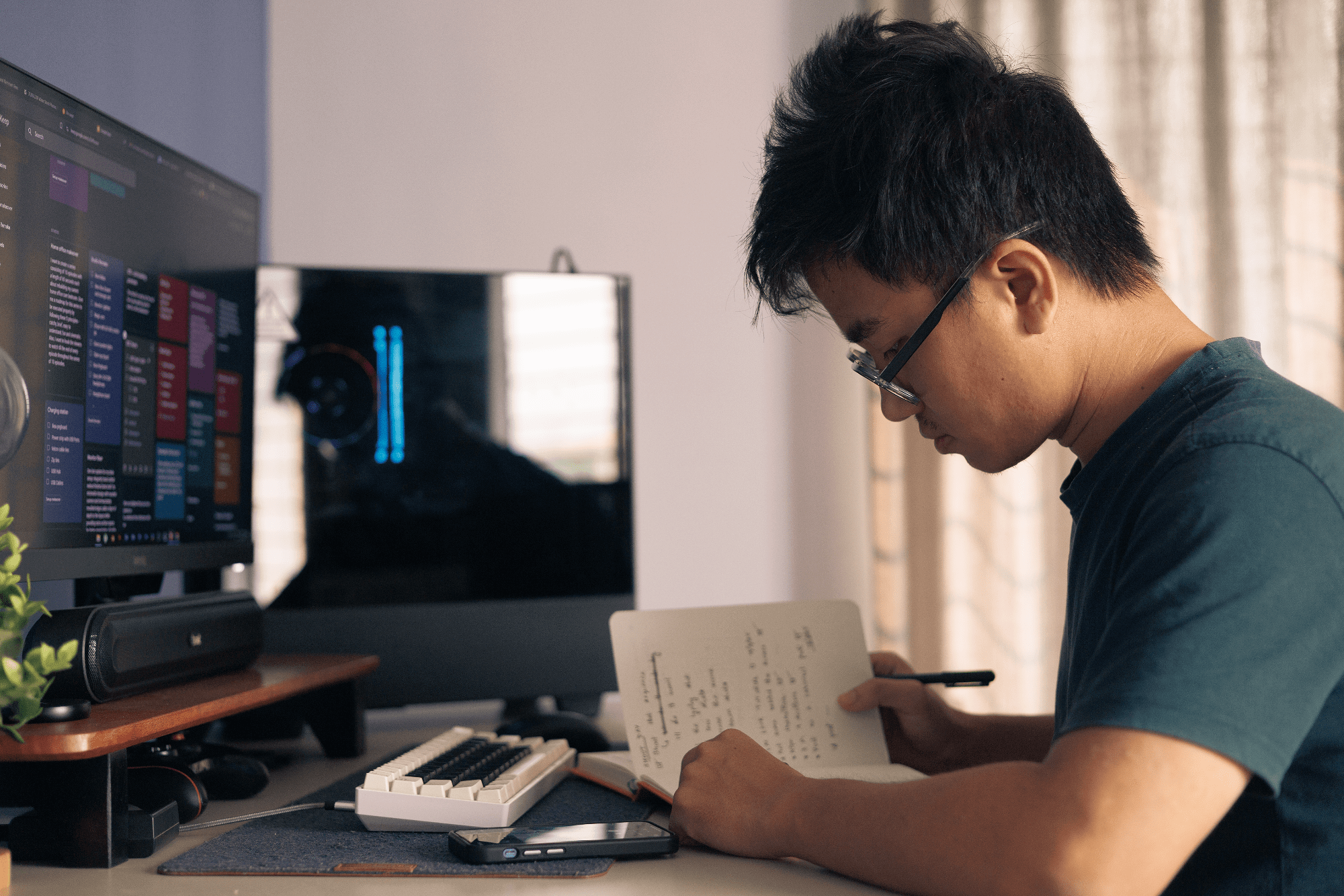 A man writing in a notebook at a home office desk with a BenQ PD2500Q monitor and an EPOMAKER Skyloong SK61 keyboard