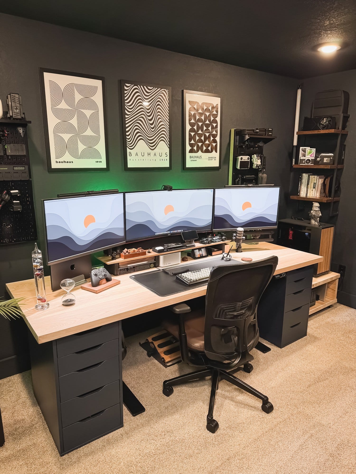 Modern desk setup with triple monitors, ergonomic chair, wooden desk with drawers, wall-mounted accessories, custom PC with green lighting, and minimalist decor in a home office