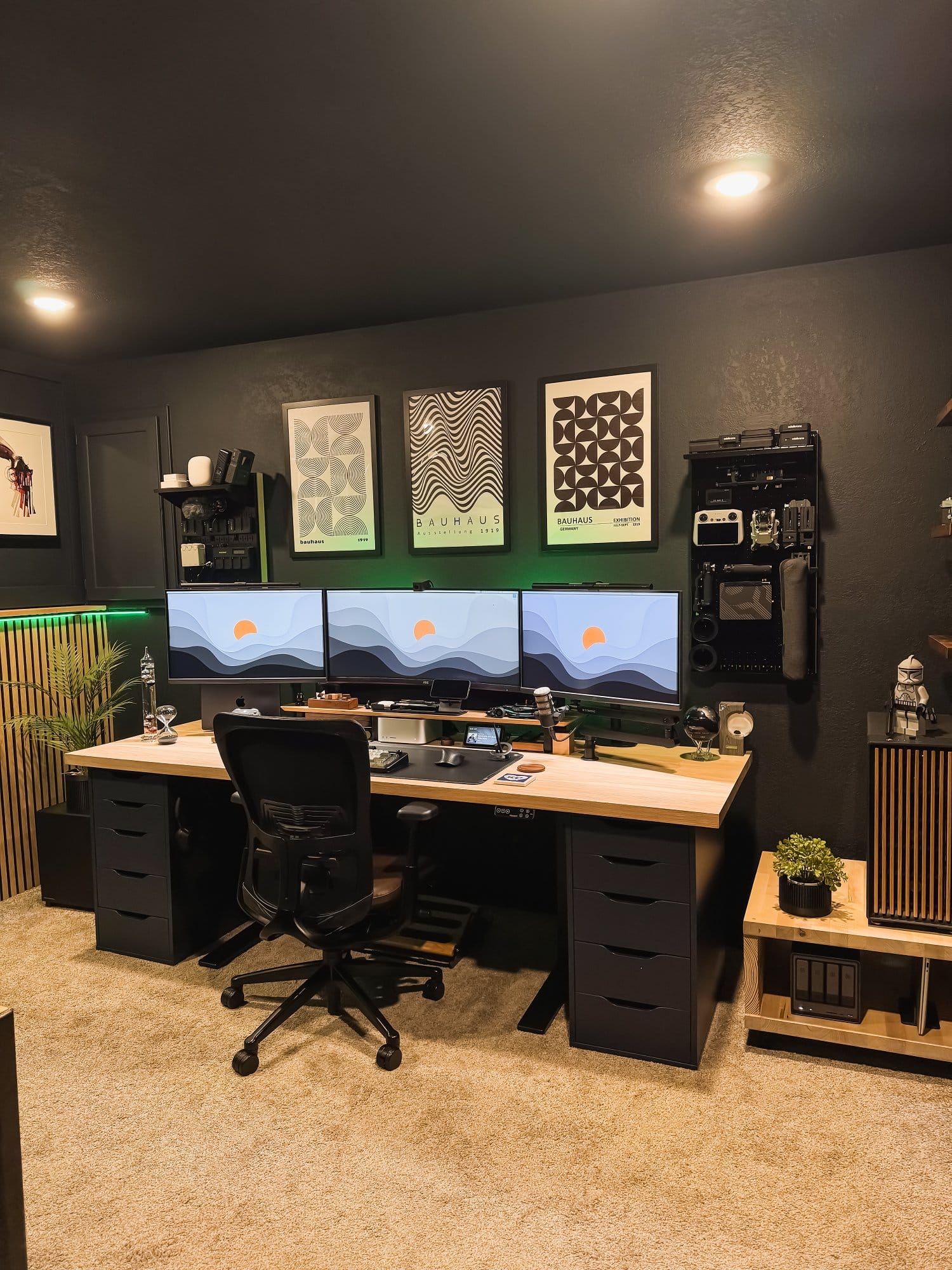 modern home office desk setup with triple monitor display, ergonomic chair, wooden desk with black drawer units, wall art decor, organised gear wall panel, and ambient lighting