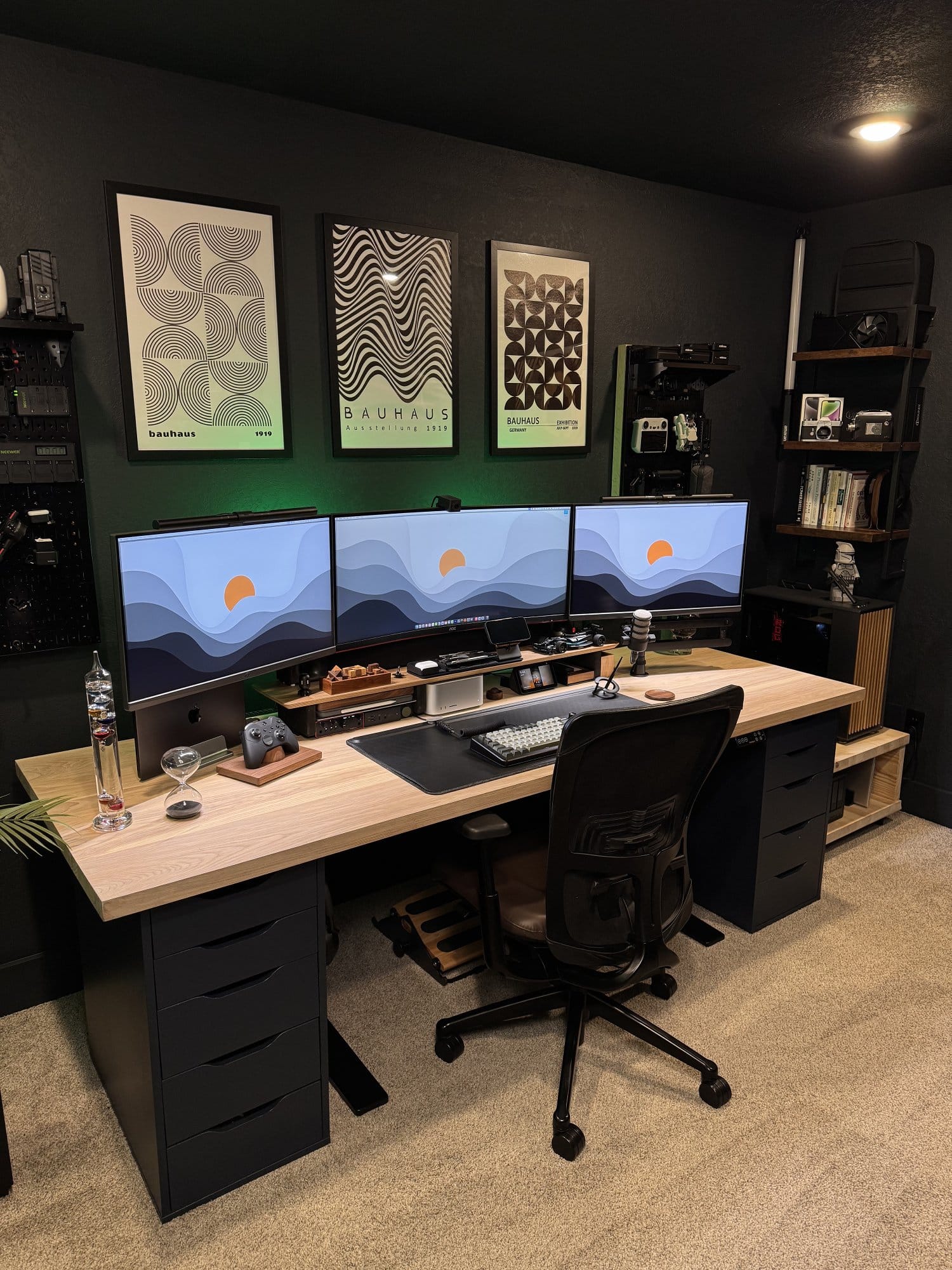 minimalist home office setup with triple monitors, wooden desk with black drawer units, ergonomic chair, wall mounted art prints, and neatly organised workspace accessories