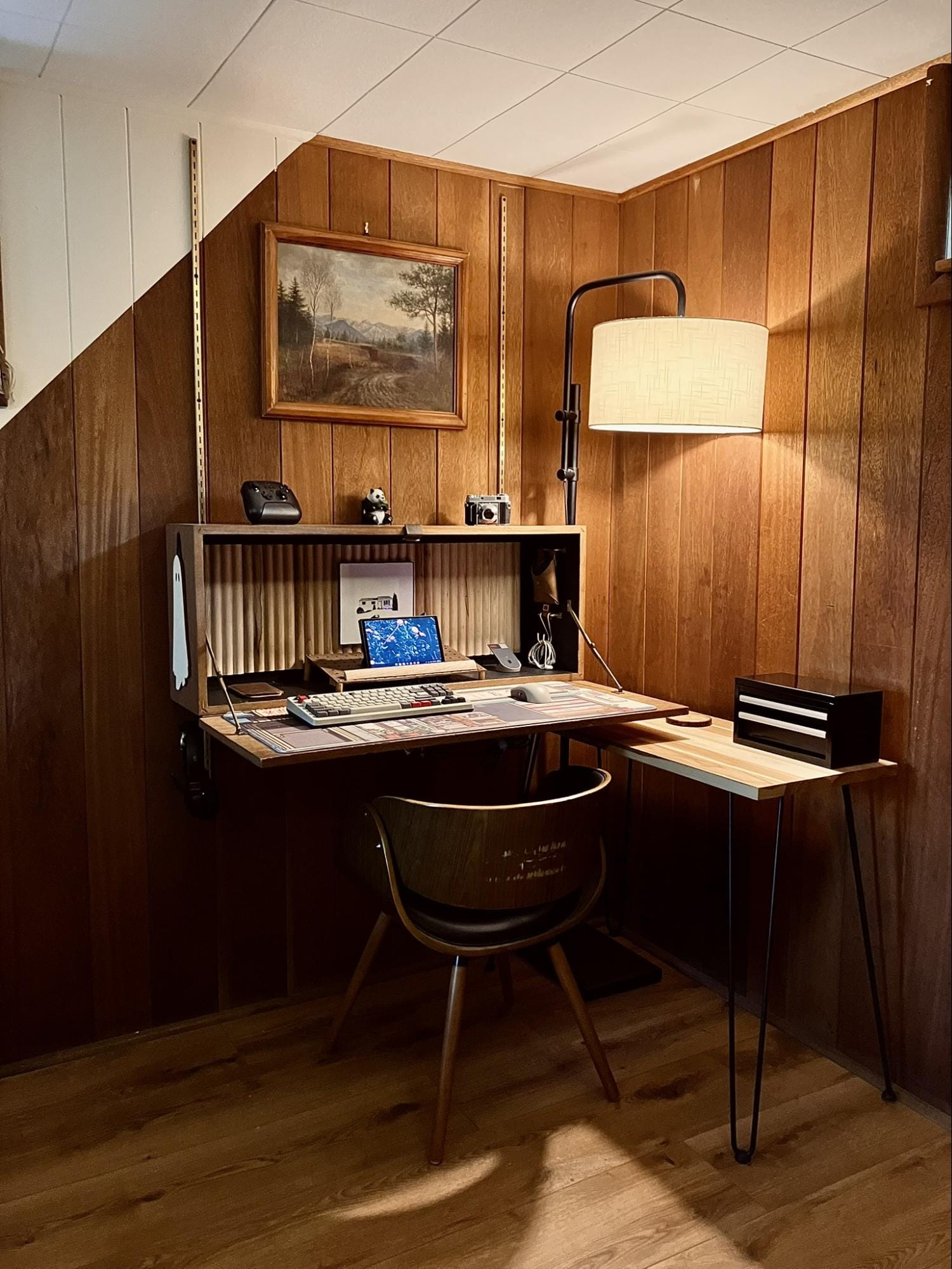 minimalist vintage desk setup with wood panel walls, floating fold-out desk, a chair, lamp, and framed painting