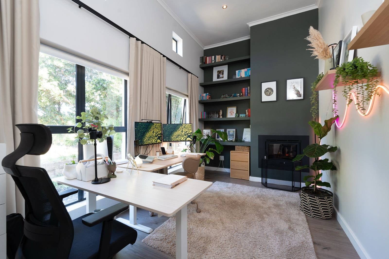 modern home office with dual monitor desk setup, ergonomic chair, large windows with natural light, green accent wall with built-in bookshelves, indoor plants, and cosy workspace décor