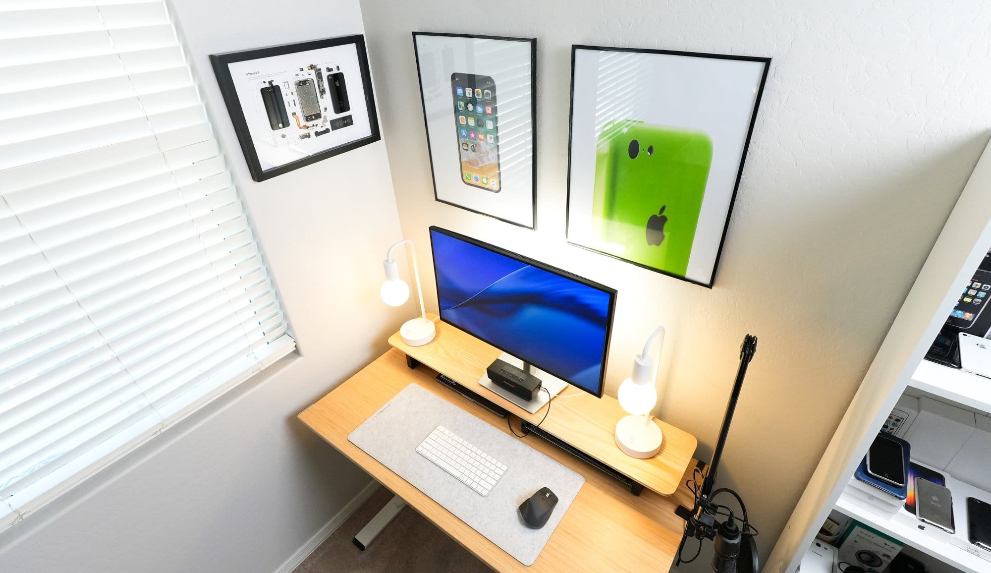 minimalist desk setup with single monitor, wireless keyboard and mouse, dual desk lamps, microphone arm, bamboo desk, and framed Apple-inspired wall art