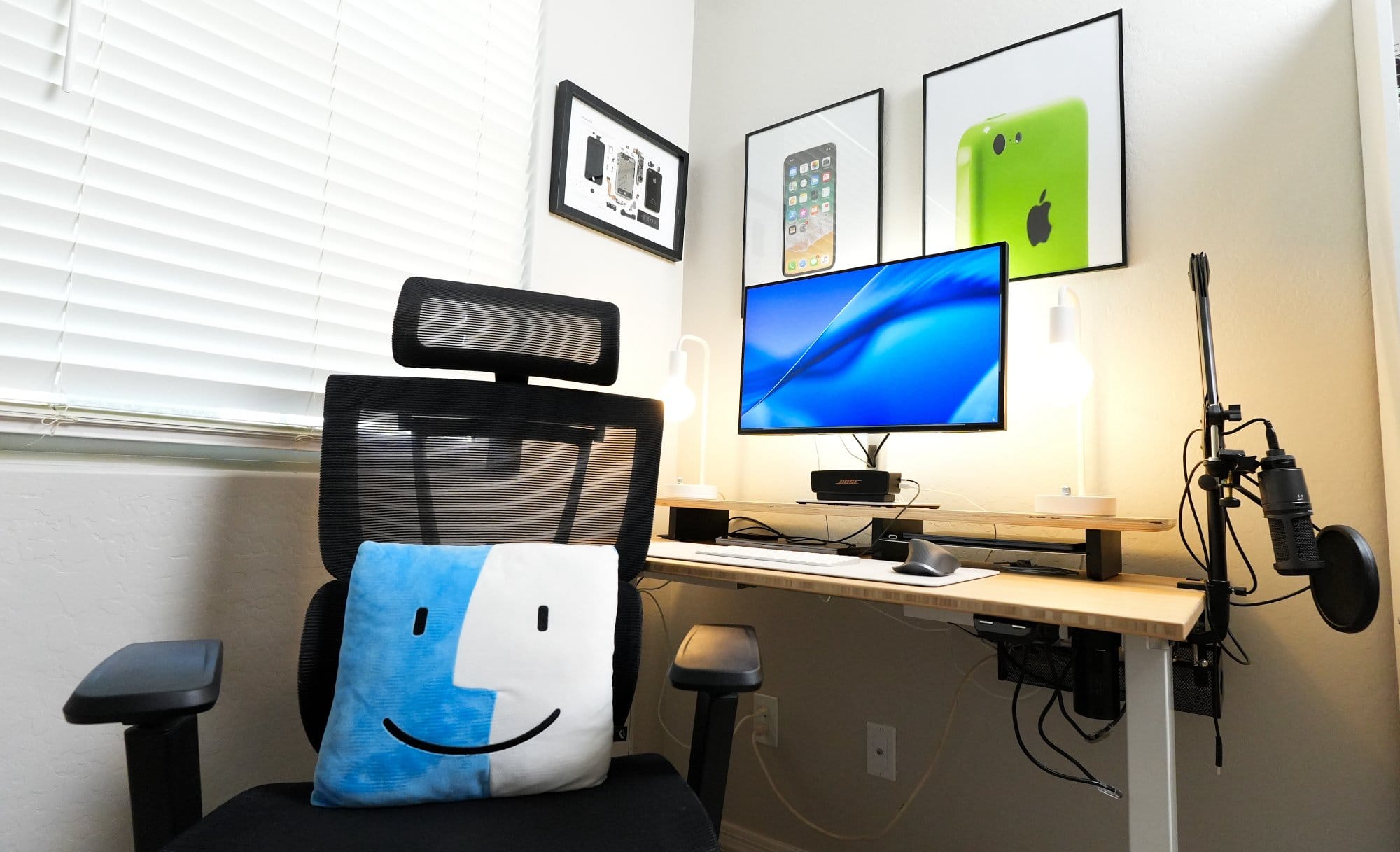 modern home office desk setup with ergonomic chair, single monitor, wireless keyboard and mouse, dual desk lamps, microphone arm, bamboo desk, and wall art featuring deconstructed Apple iPhone and classic iPhone prints