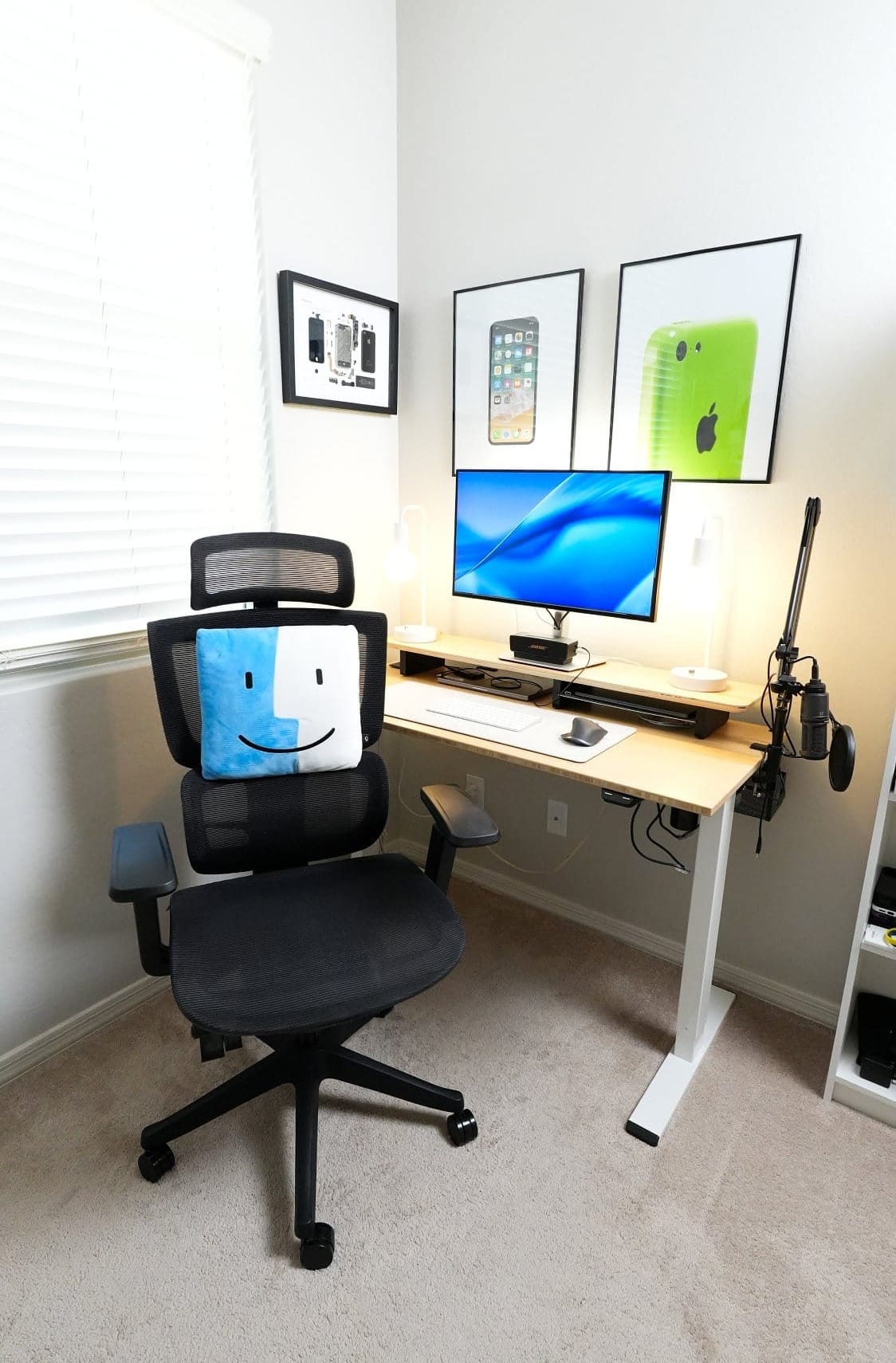 ergonomic home office desk setup with mesh chair, Apple Finder pillow, single monitor, wireless keyboard and mouse, dual desk lamps, microphone arm, standing desk, and wall art featuring a deconstructed Apple iPhone and iPhone prints