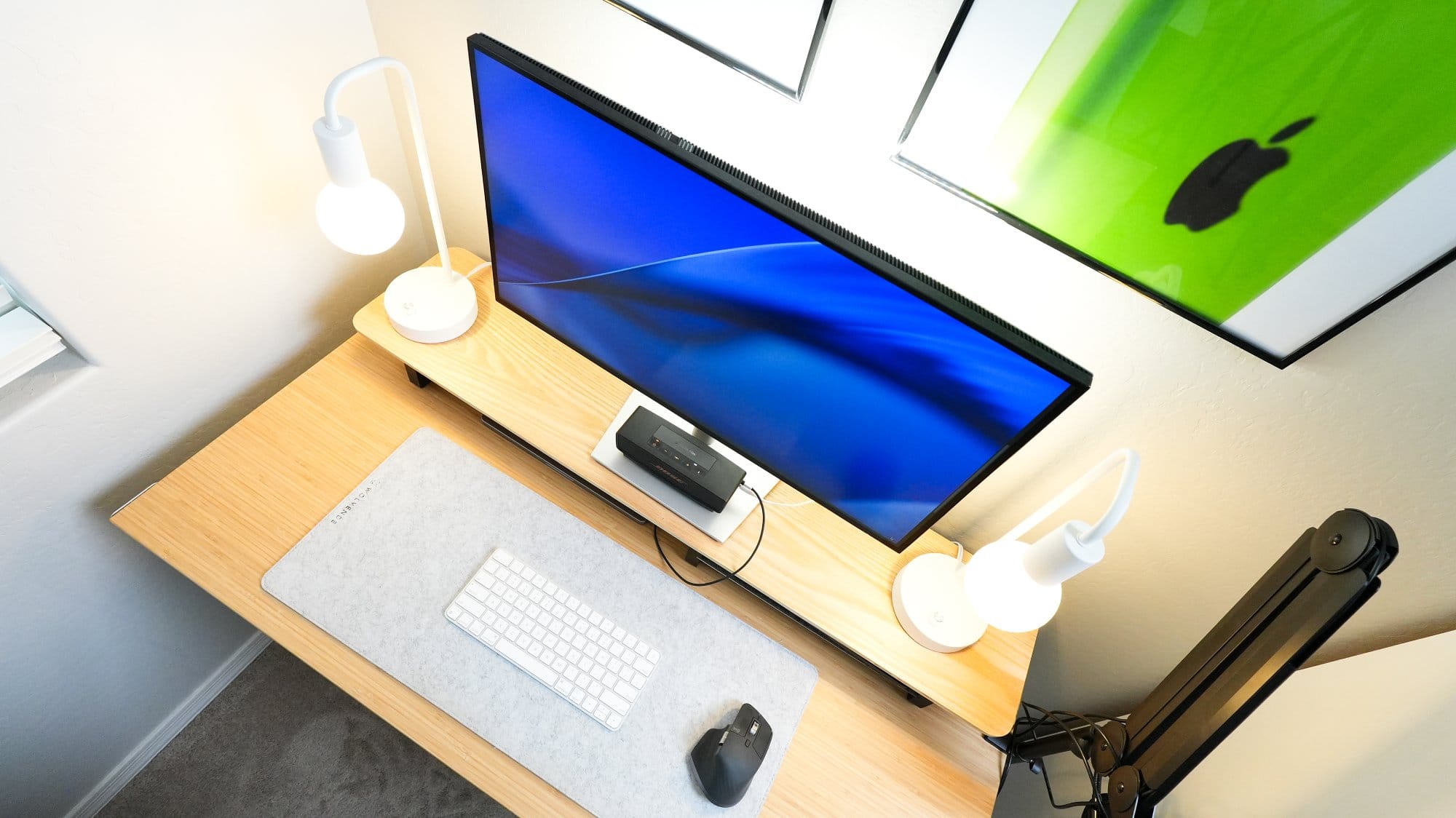 top view of minimalist desk setup with single monitor, wireless keyboard and mouse, dual desk lamps, bamboo desk, and framed Apple artwork on the wall
