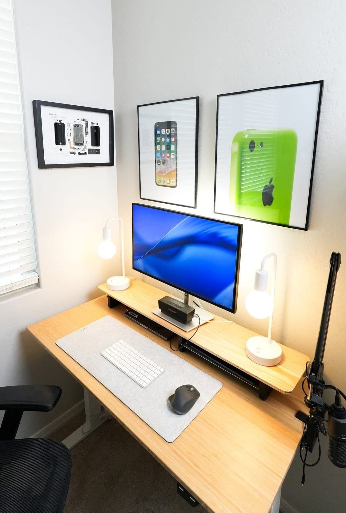 minimalist desk setup with single monitor, wireless keyboard and mouse, dual desk lamps, microphone arm, bamboo desk, and framed Apple-inspired wall art