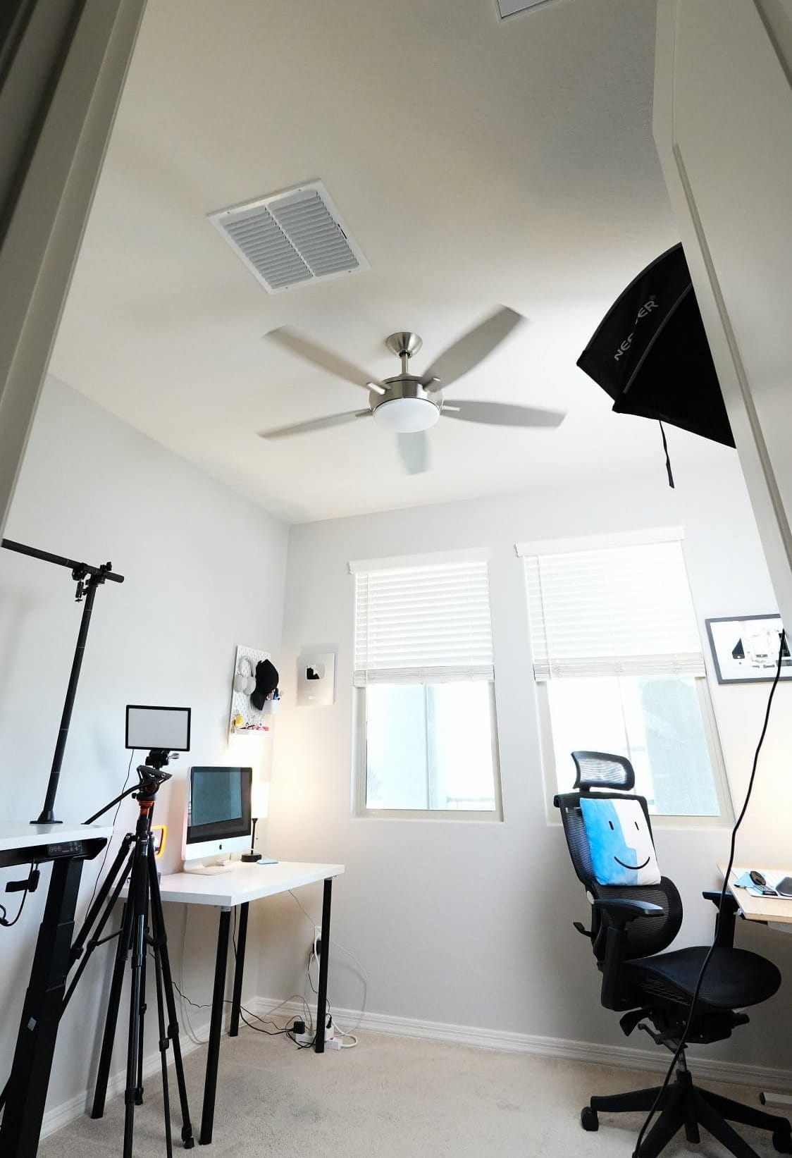 overview of modern content creator workspace with dual desk setups, iMac workstation, ergonomic chair, overhead camera rig, tripod lighting, and wall-mounted gear in a bright home office