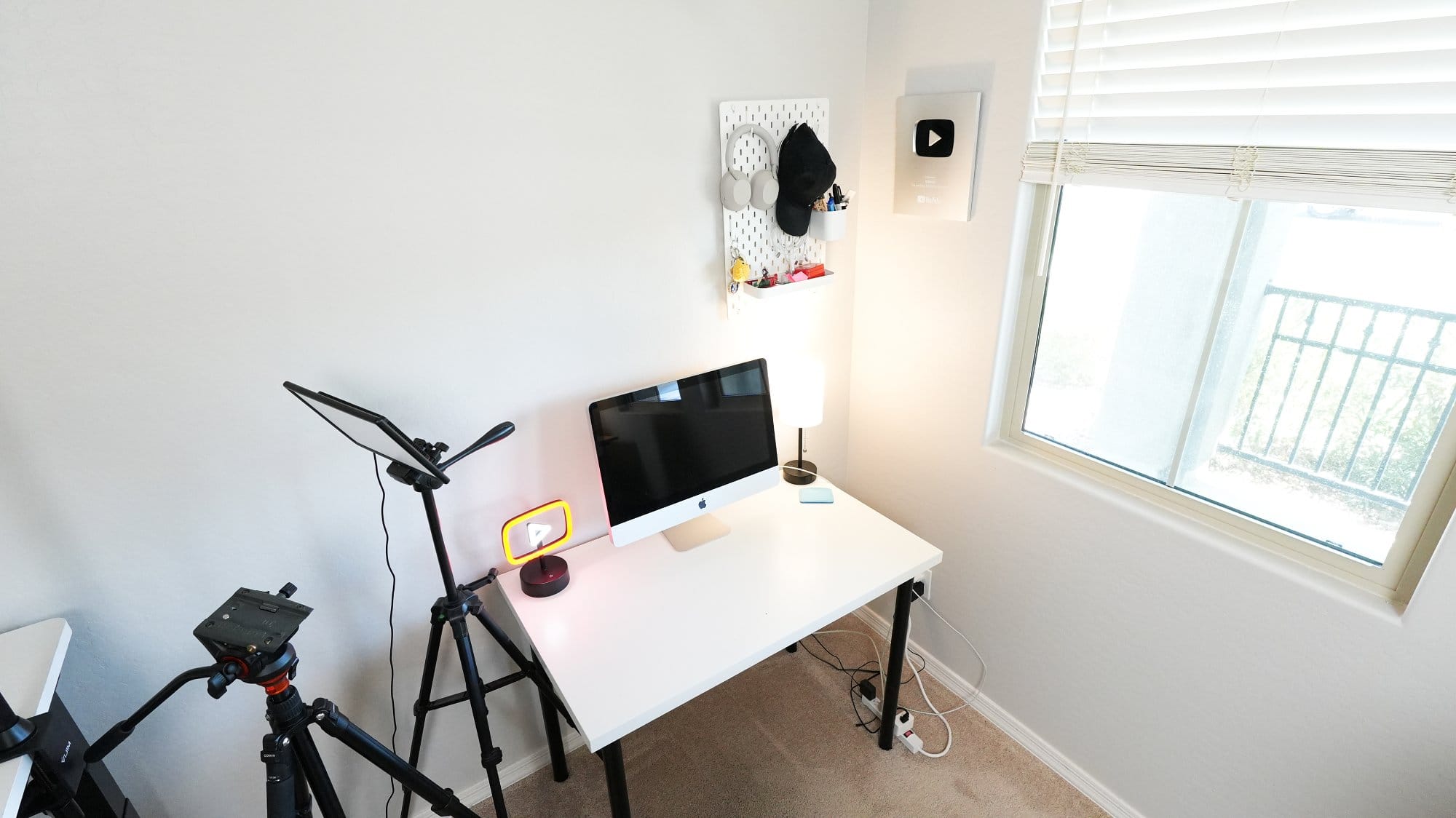 minimal white desk setup with Apple iMac, tripod lighting, desk lamp, pegboard wall organiser with headphones and accessories, and YouTube creator award