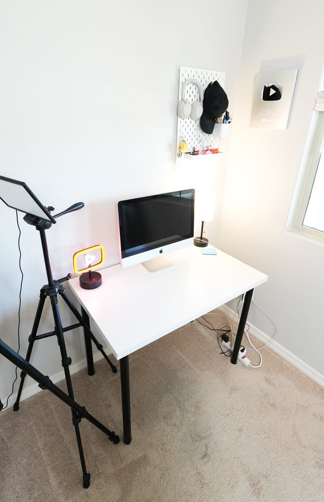 minimal white desk setup with Apple iMac, tripod lighting, desk lamp, pegboard wall organiser with headphones and accessories, and YouTube creator award