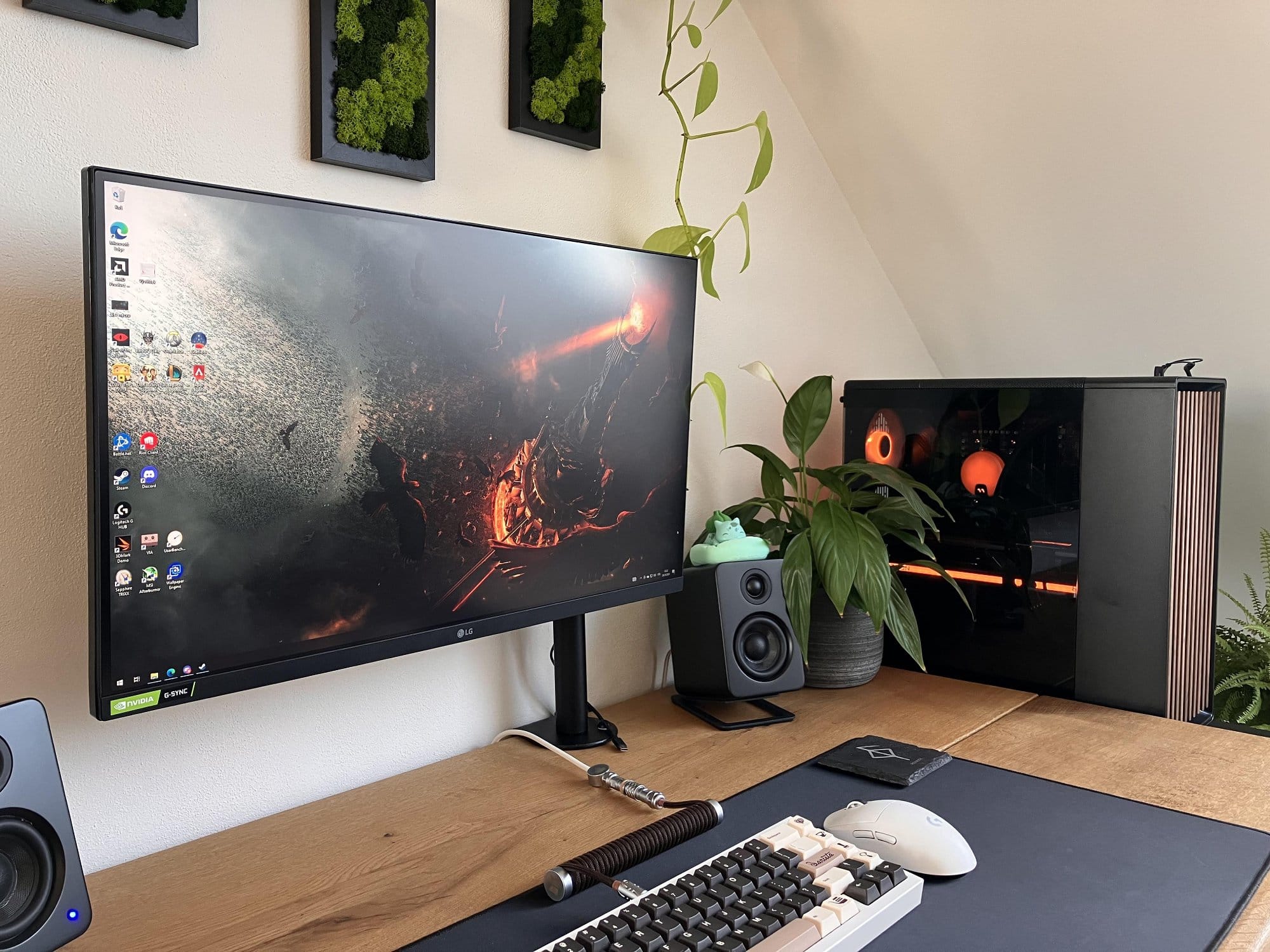 A home office desk with a large monitor displaying a fiery scene, speakers, keyboard, mouse, and a potted plant next to a sleek black computer case with orange lighting inside