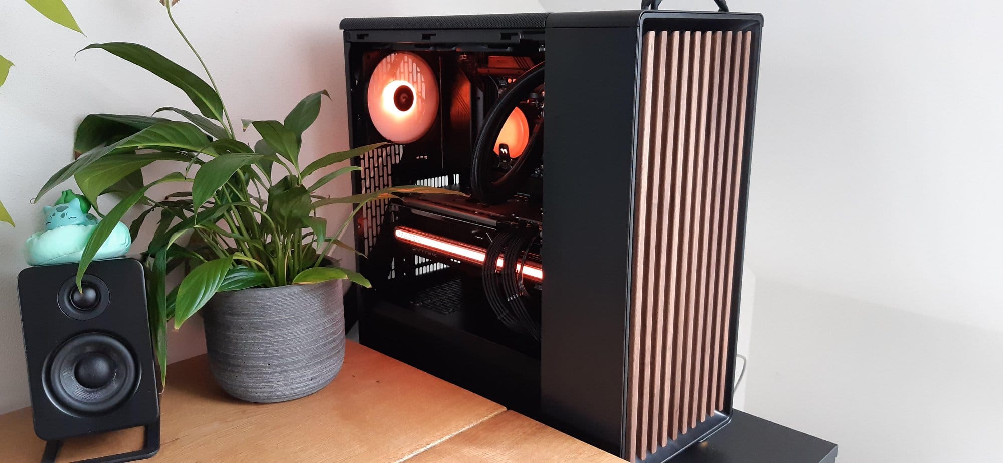 A close-up view of an open computer case with internal components illuminated by orange lights, next to a potted plant and a speaker adorned with a small figurine