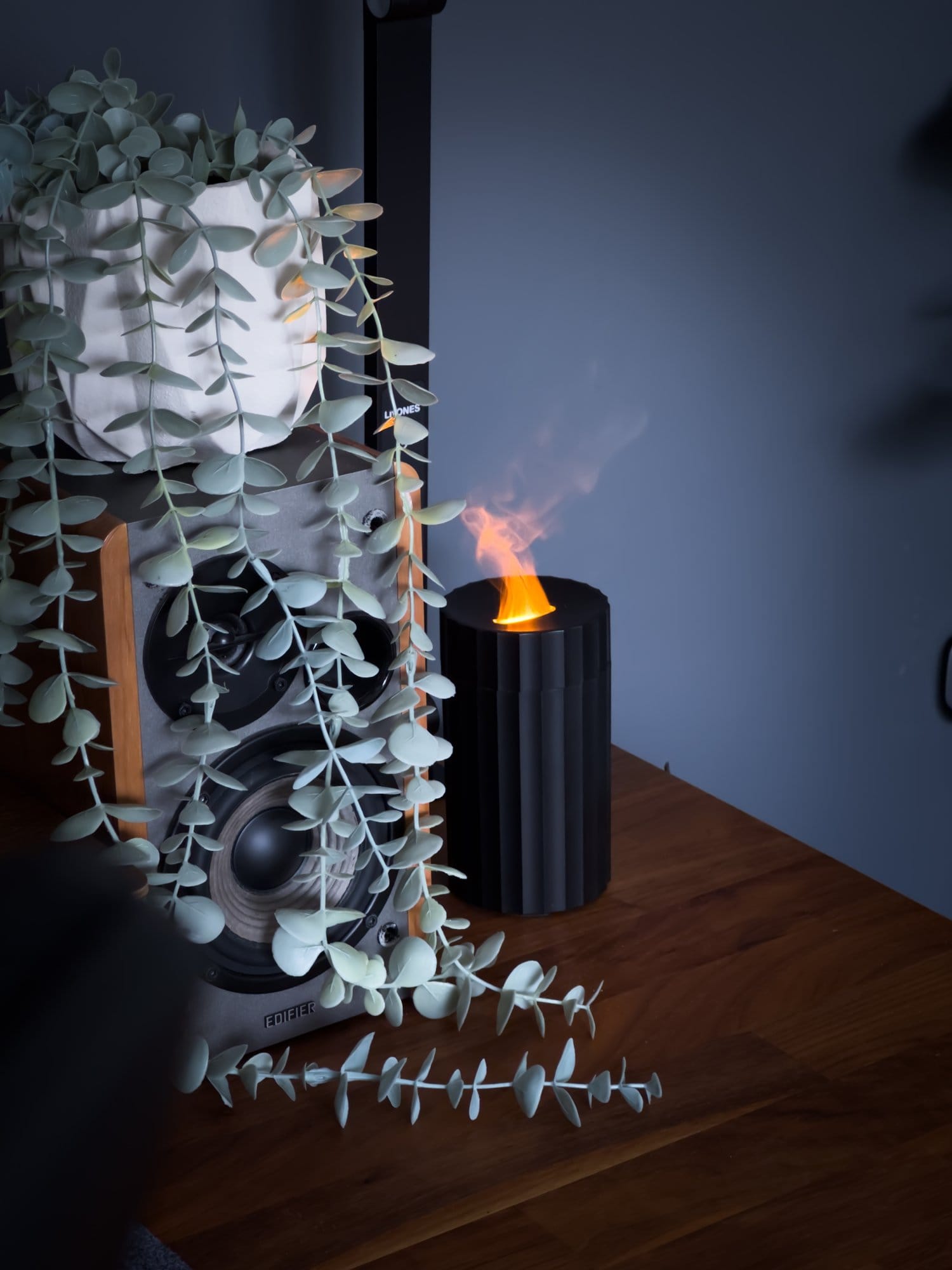 A desk setup featuring an Edifier R1280T speaker adorned with a trailing plant and an MDS Candela Diffuser emitting a warm, flame-like glow