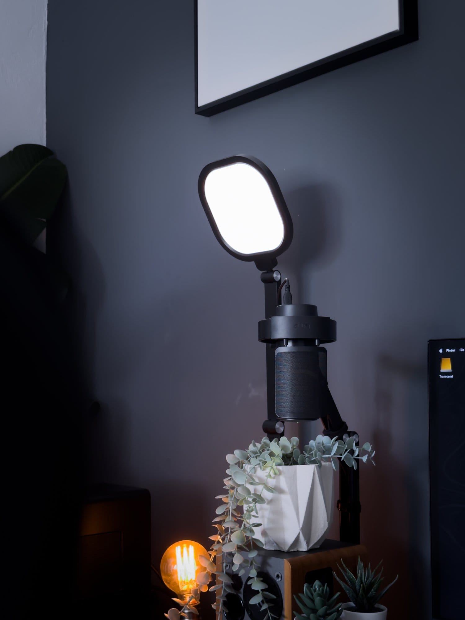 A desk setup featuring a LitONES Square 3 Plus light, a Fifine AM-8 microphone on a Ku Xiu X36 boom arm, and a vintage-style bulb lamp next to a potted plant