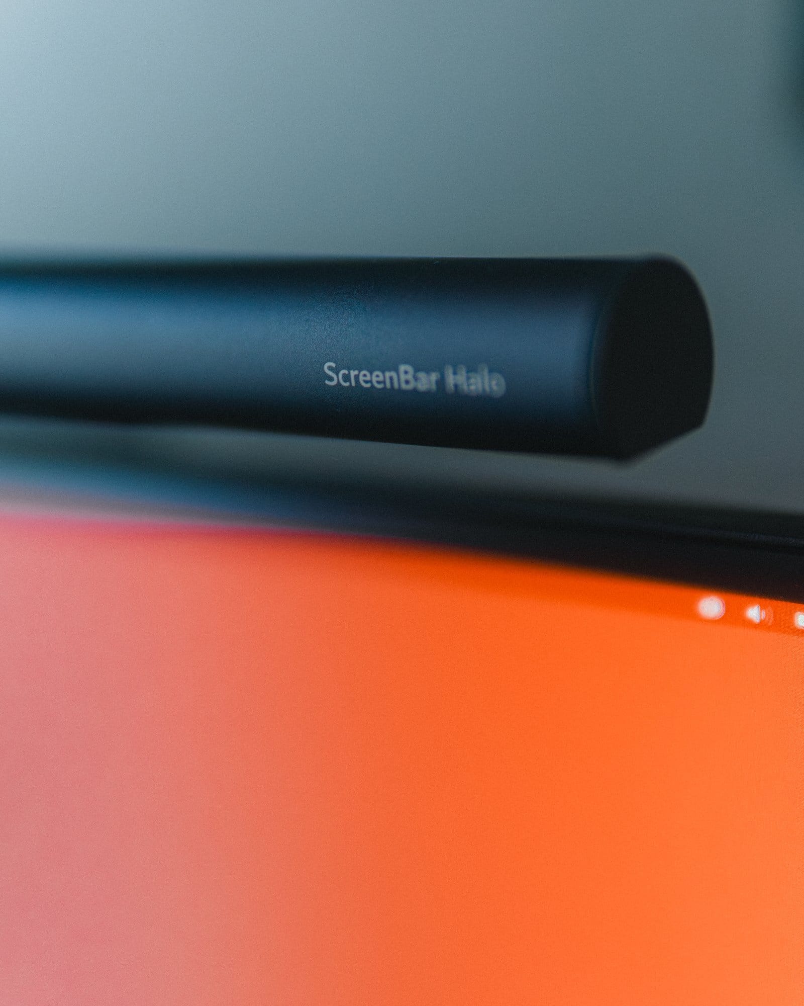 A close-up of a BenQ ScreenBar Halo light bar mounted on the top edge of a monitor, with the screen showing a blurred orange display in the background