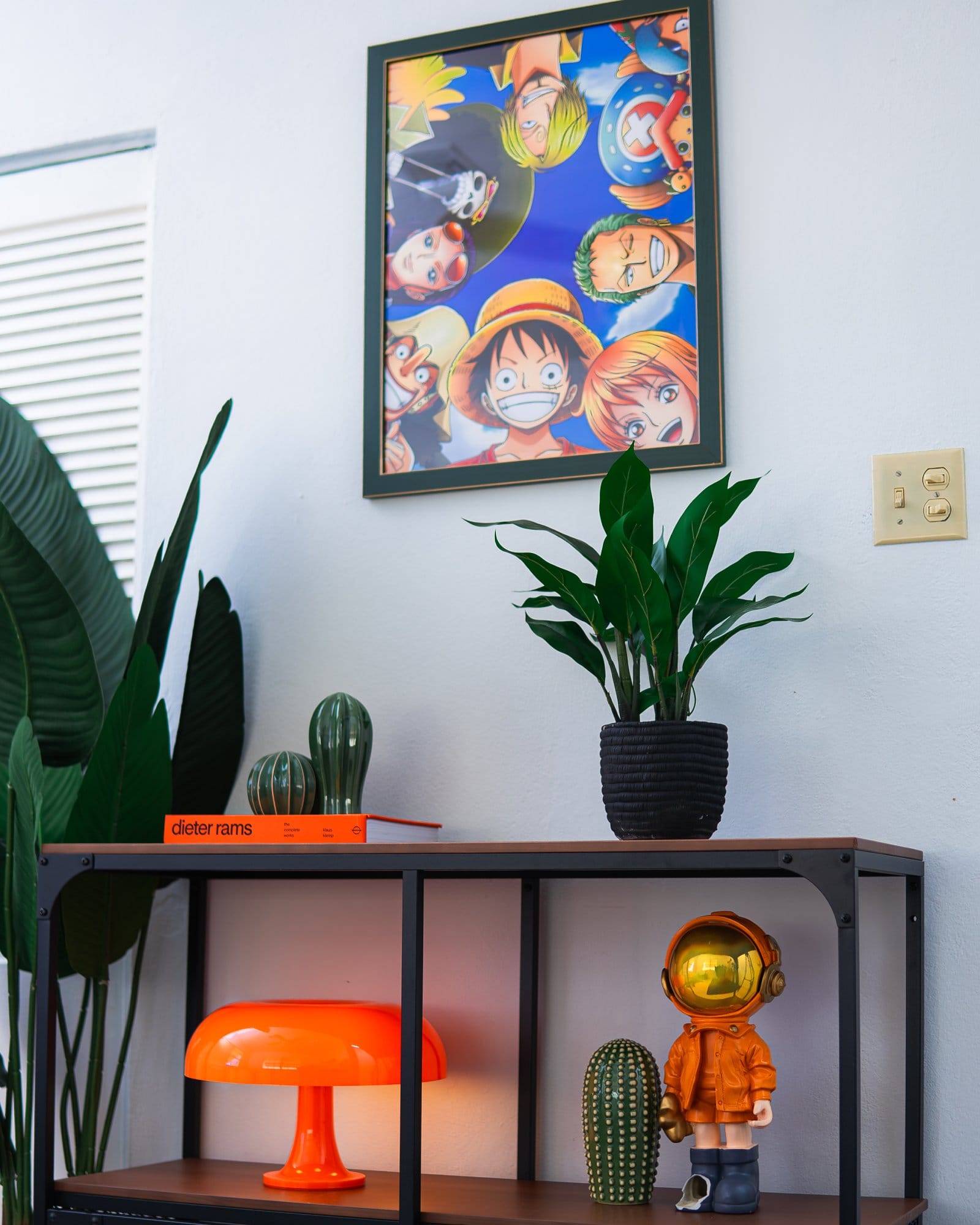 A stylish shelving unit featuring an orange mushroom lamp, a white speaker, an orange astronaut figurine, green cactus decor pieces, an orange Dieter Rams book, and a black potted plant, with a One Piece anime poster mounted on the wall above