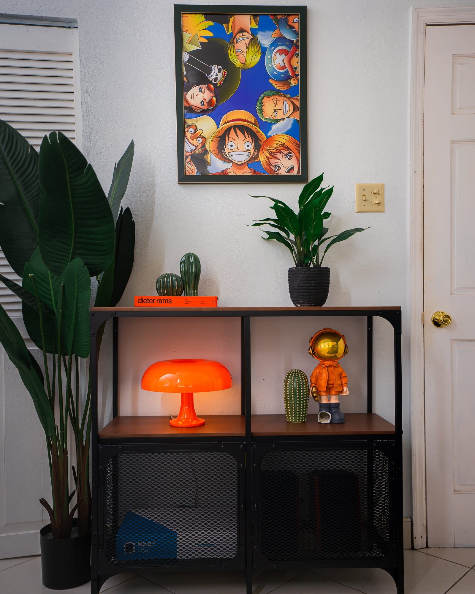 A well-decorated shelf setup featuring a One Piece anime poster on the wall, a tall green plant to the left, a Dieter Rams book, a small green cactus decor piece, an orange mushroom lamp, and a stylised astronaut figurine in an orange suit with a reflective gold visor