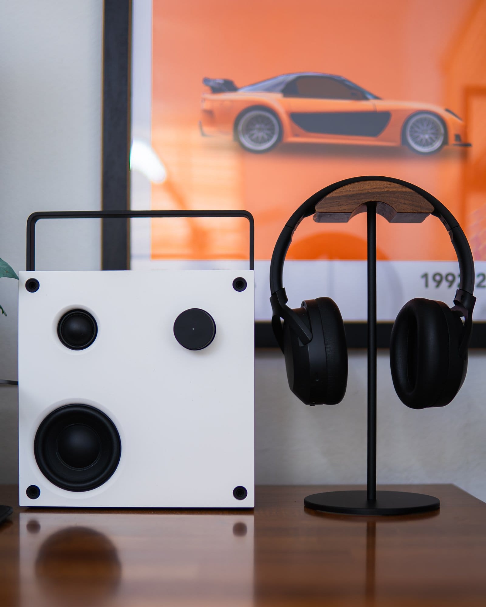 A stylish desk setup featuring a minimalist white speaker, black headphones on a stand, and an orange poster of a sports car in the background