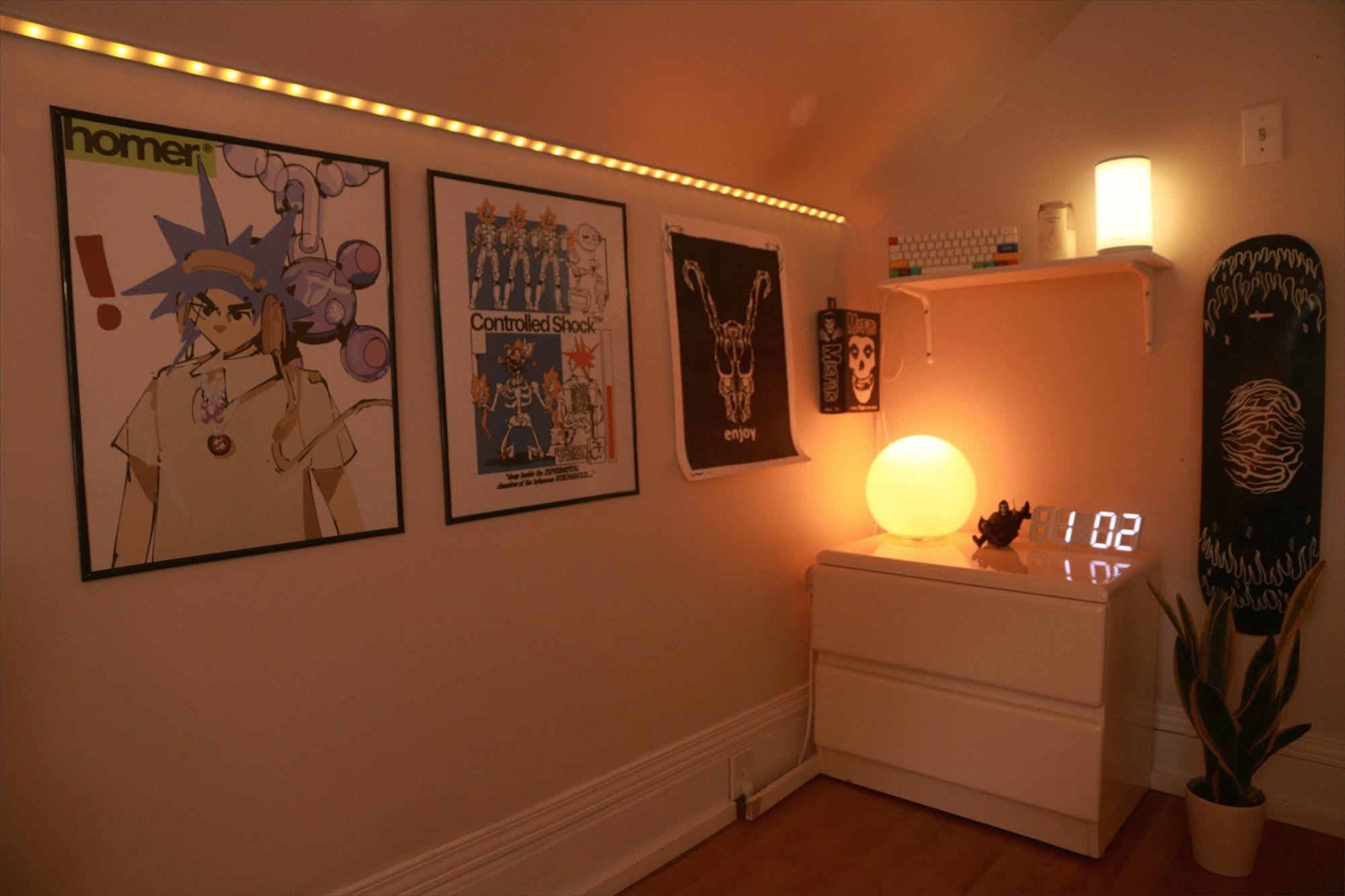 A stylish corner with framed posters, a digital clock, and ambient lighting, featuring a white chest of drawers and a modern globe lamp, complemented by a potted plant and a skateboard deck on the wall
