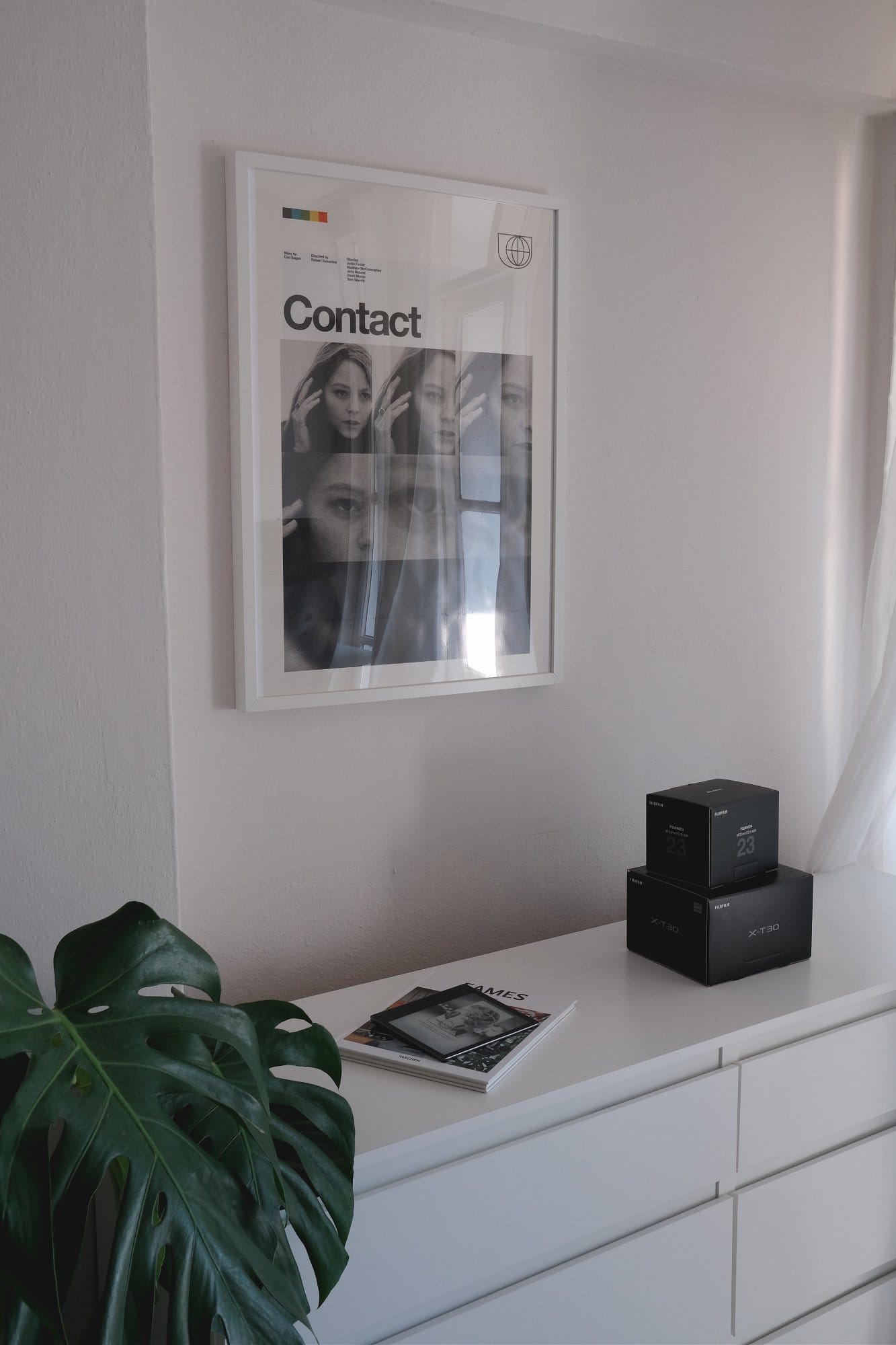A framed poster titled “Contact” hangs above a white dresser with Fujifilm X-T30II camera boxes and books on top, alongside a large leafy Monstera plant