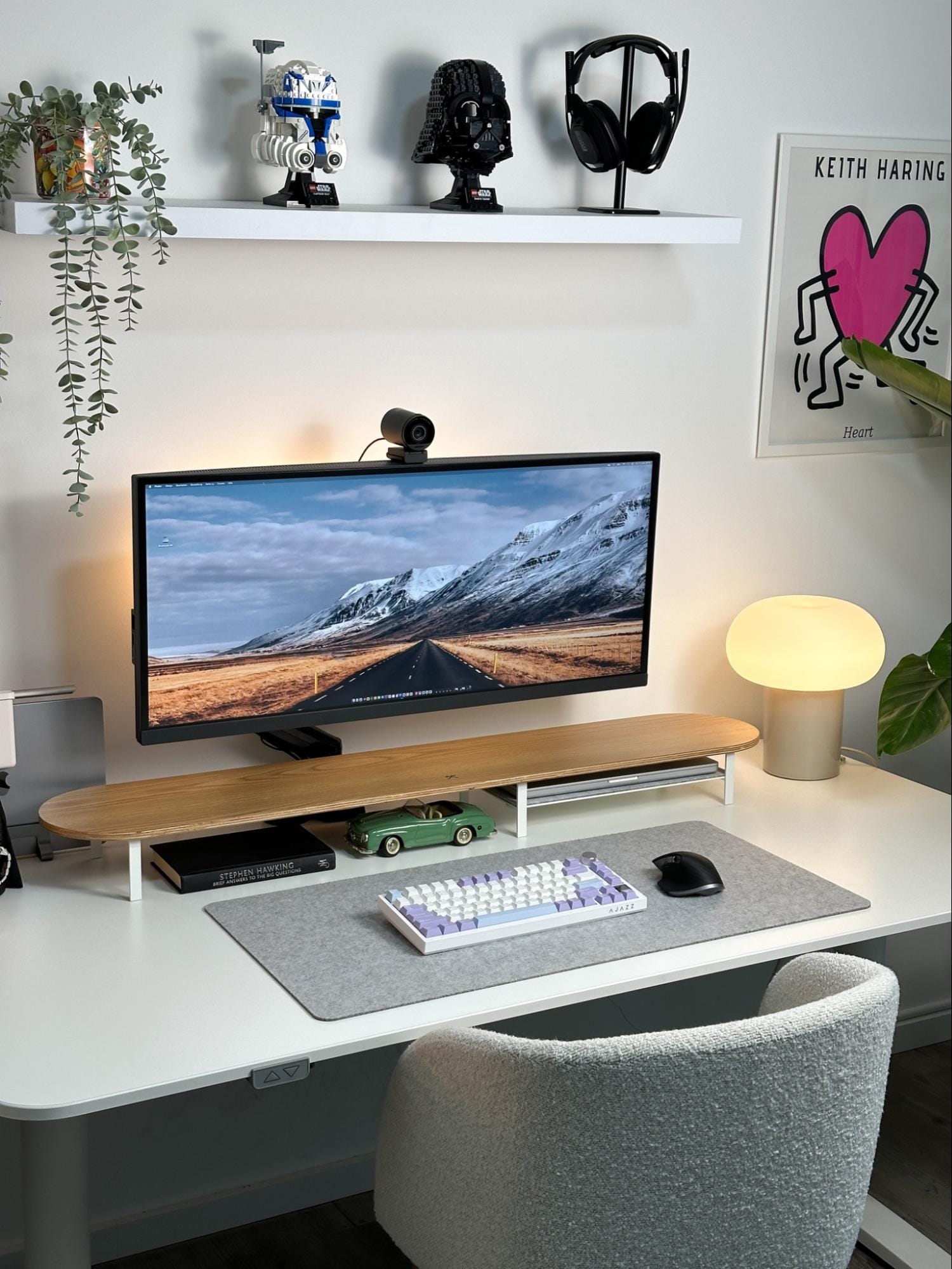 A well-organised workspace featuring a BenQ monitor, a mechanical keyboard, and a Logitech MX Master 3S mouse on a felt mat, a Woodcessories desk shelf, a green toy car, a book by Stephen Hawking, and a modern lamp, with LEGO Star Wars Darth Vader and Captain Rex helmets and an Astro A50 headset on a shelf above, and a Keith Haring “Heart” poster on the wall
