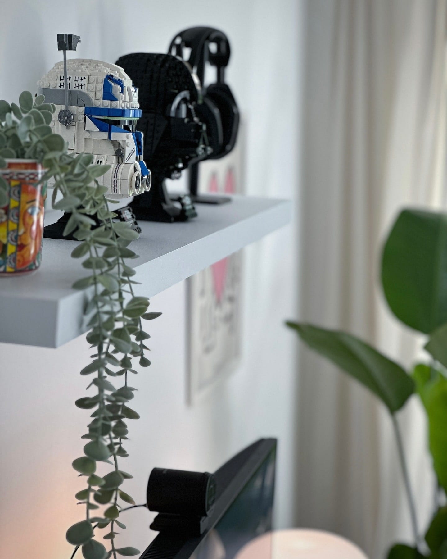 A close-up of a shelf displaying LEGO Star Wars Captain Rex and Darth Vader helmets, an Astro A50 headset, and a potted plant, with a Keith Haring “Heart“ poster visible in the background