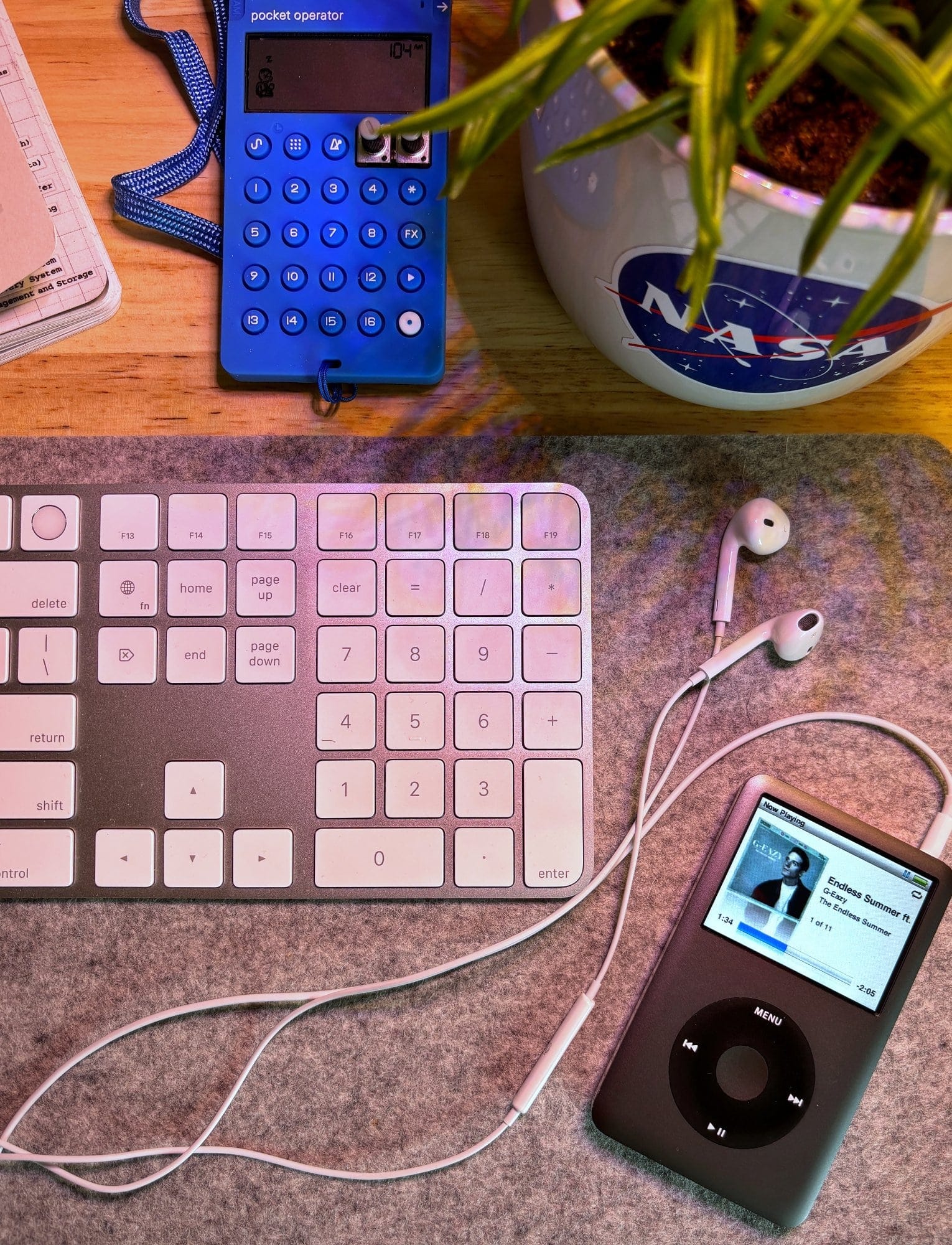 A close-up of a desk setup with an Apple Magic Keyboard, a black iPod Classic with white earbuds, a Teenage Engineering Pocket Operator, and a NASA-branded plant pot on a wooden surface