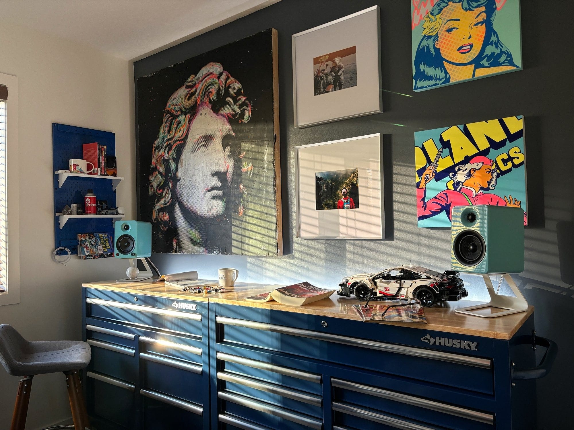 A workspace with a Husky workbench, Kanto YU4 speakers in gloss teal, a LEGO Porsche model, and framed artwork on the walls