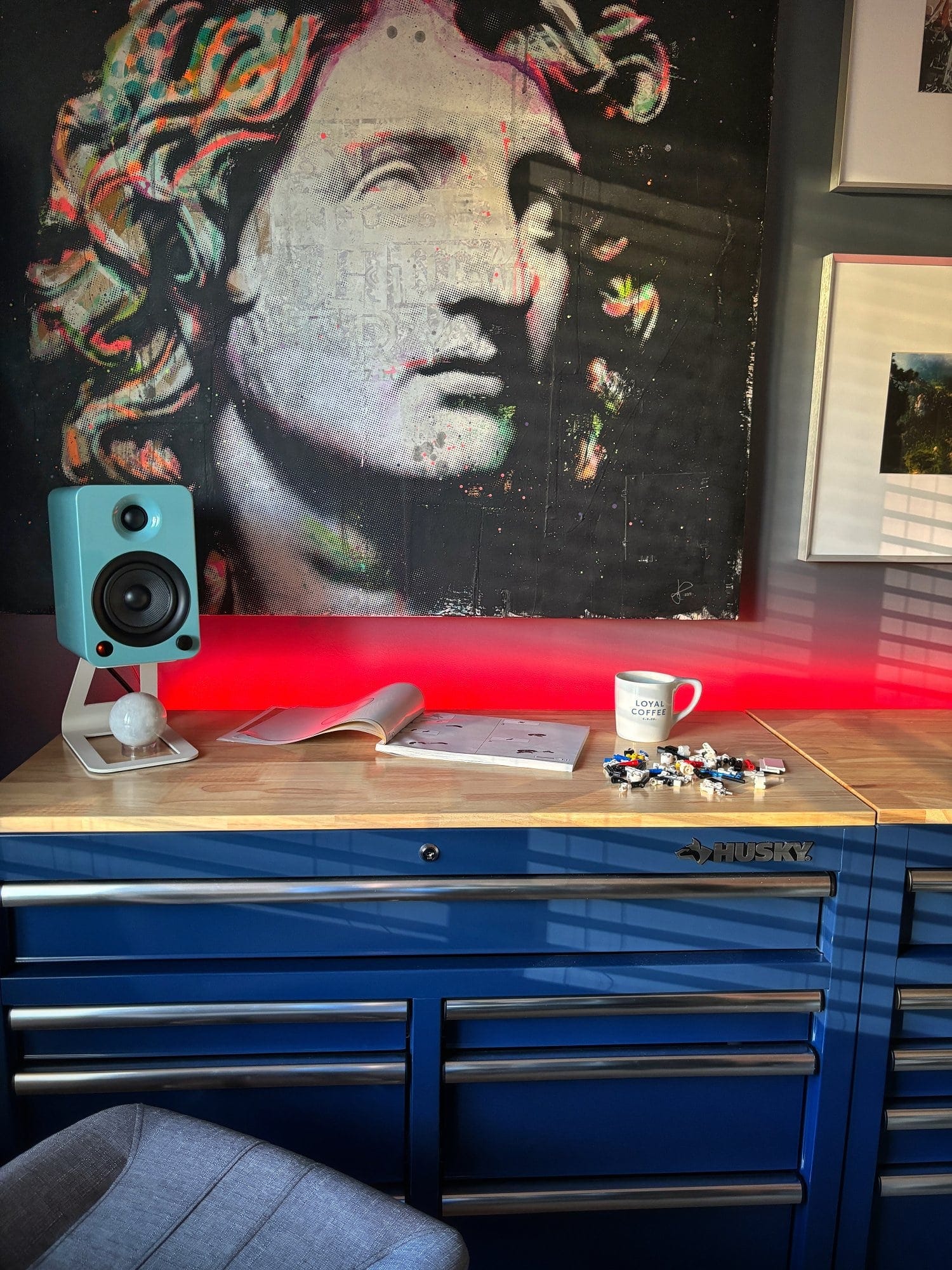 A Husky workbench with a Kanto YU4 speaker, a large sculpture print, a coffee mug, an open book, and scattered LEGO pieces, lit by red LED lighting