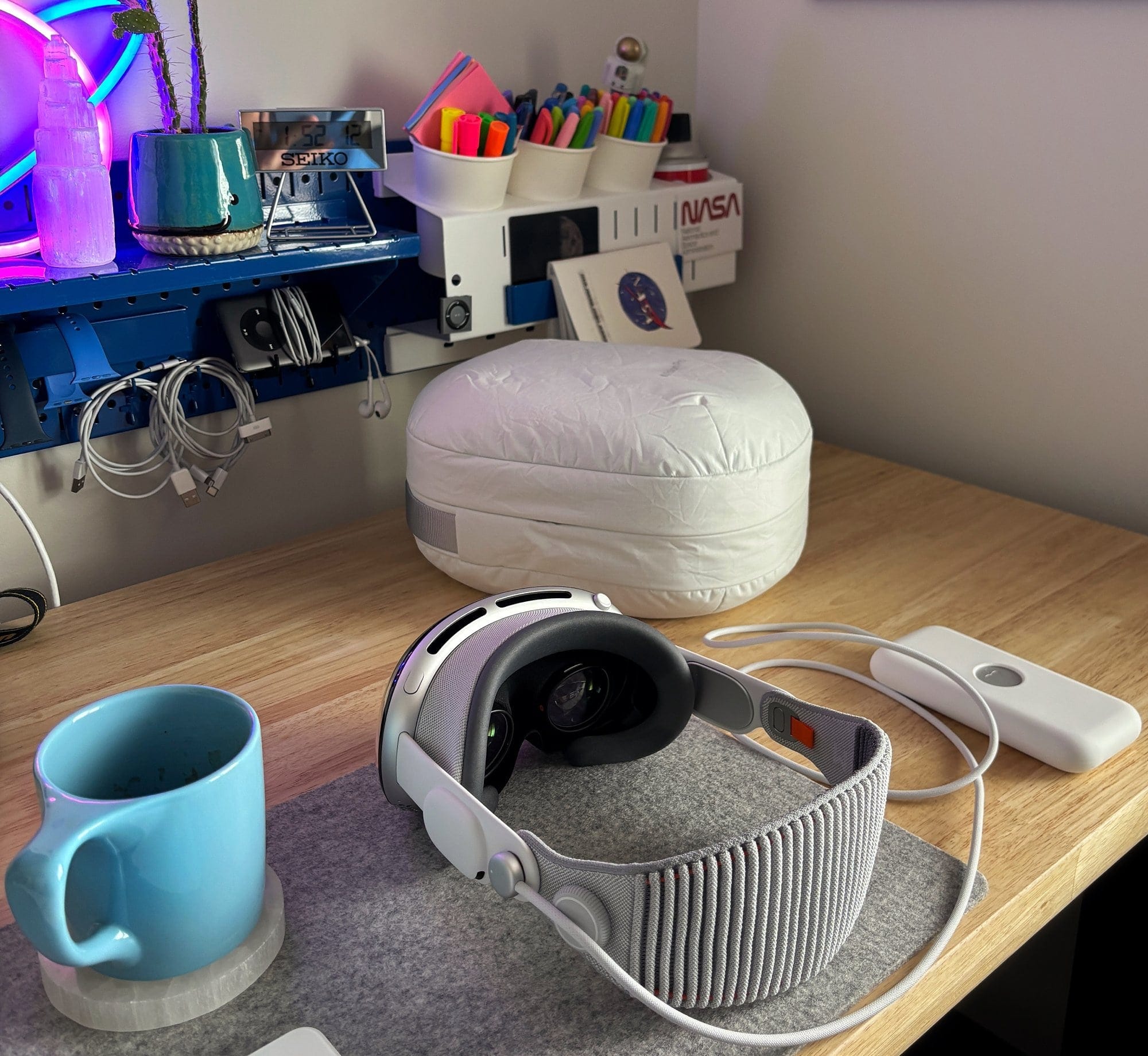 A desk setup with an Apple Vision Pro headset, its battery pack, a blue mug on a coaster, and a pen organiser mounted on the wall