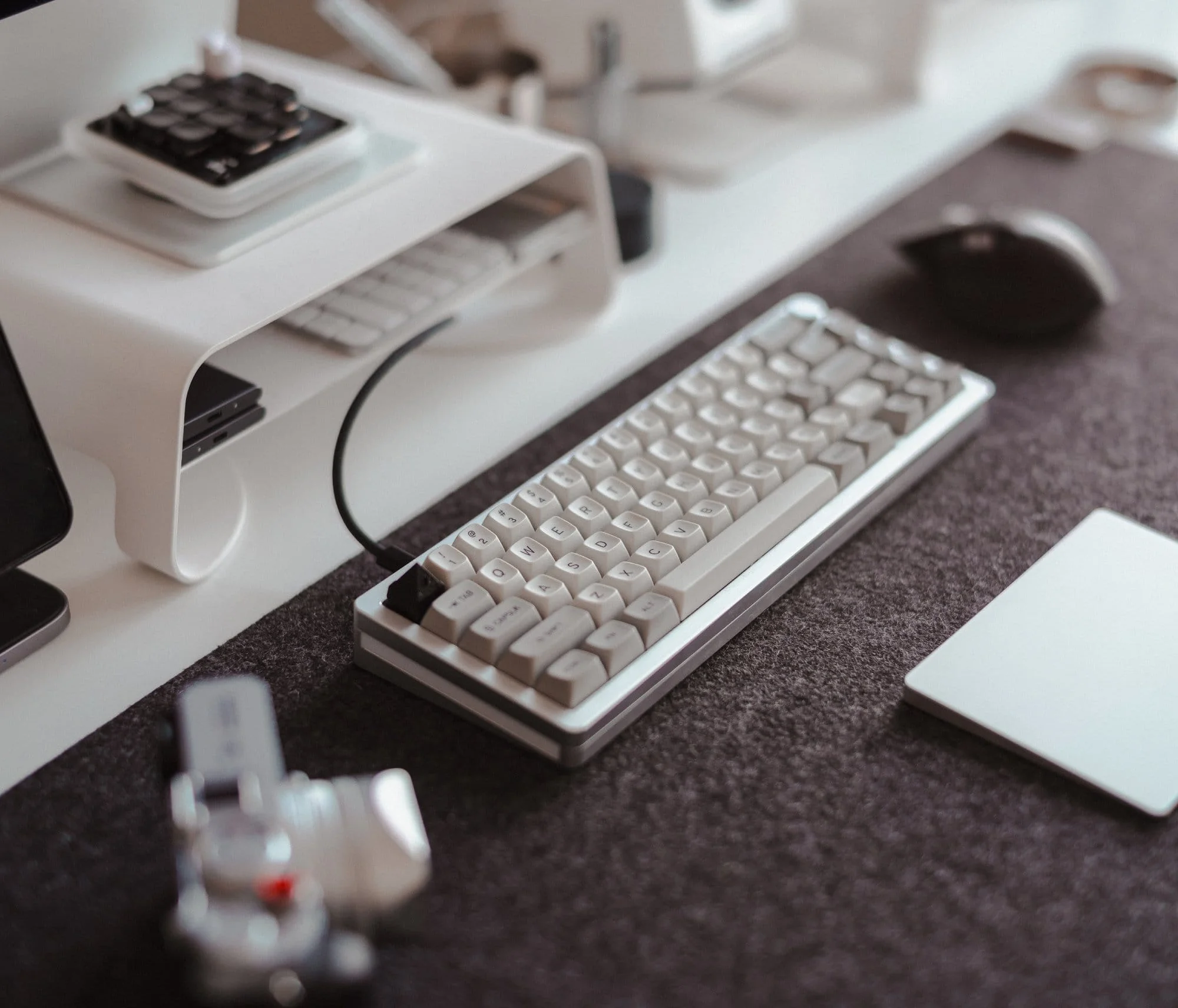 A close-up of a custom mechanical keyboard and a Logitech MX Master 3S mouse on a Grovemade desk pad, with a desk shelf above holding a numpad and wireless keyboard