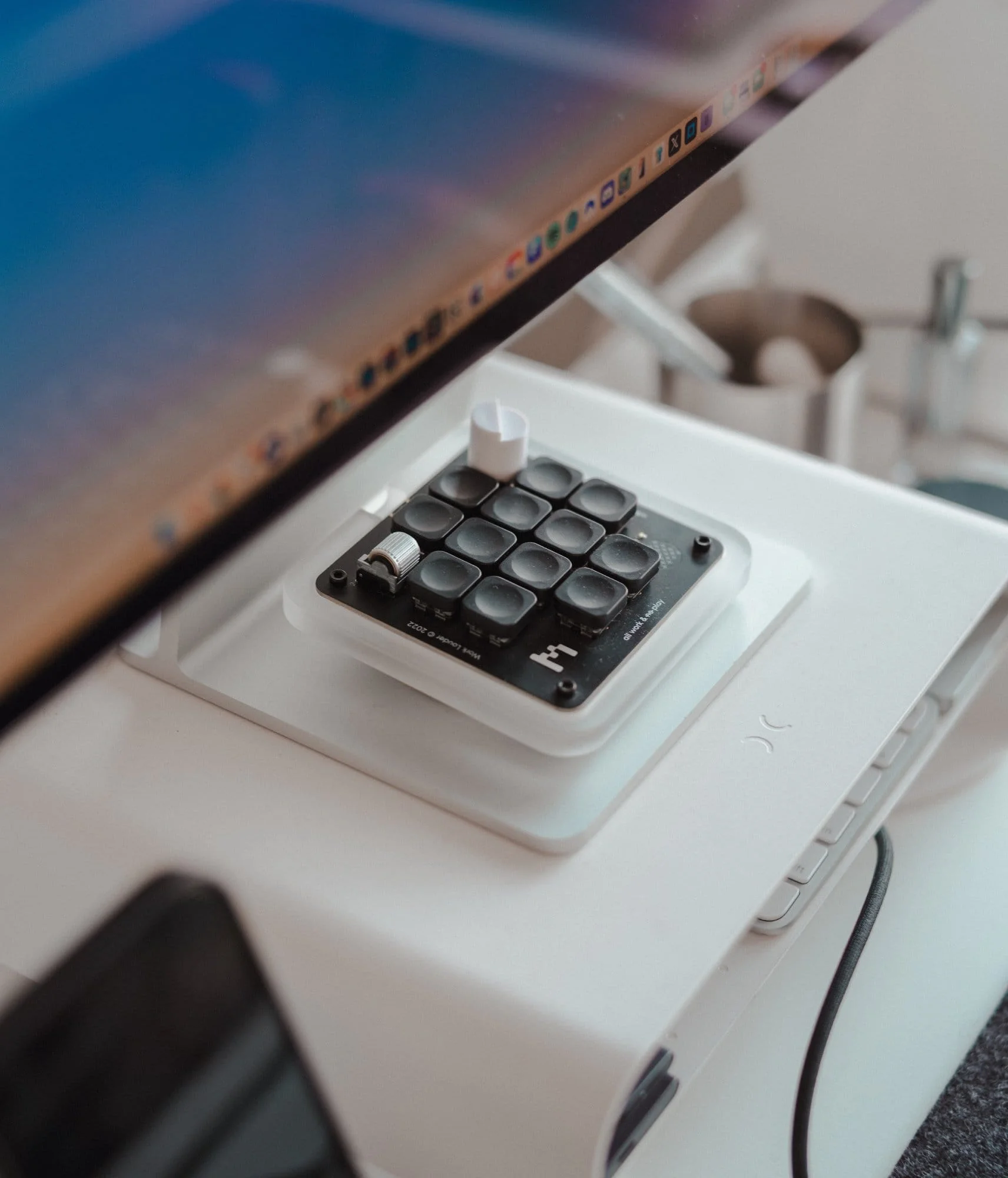 A close-up of a macro pad with dials and programmable keys sitting on a Twelve South Curve Riser, beneath an Apple Studio Display