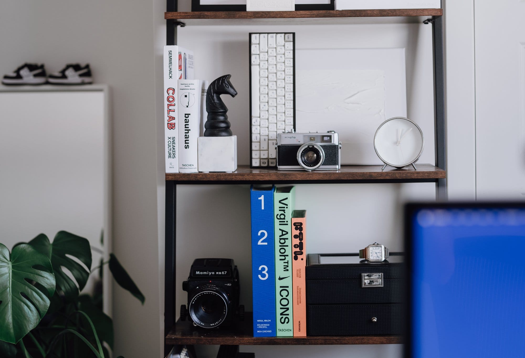 A shelf with design books, a mechanical keyboard, a Mamiya RB67 camera, a vintage Minolta camera, a small white clock, and a black storage box with a silver watch on top