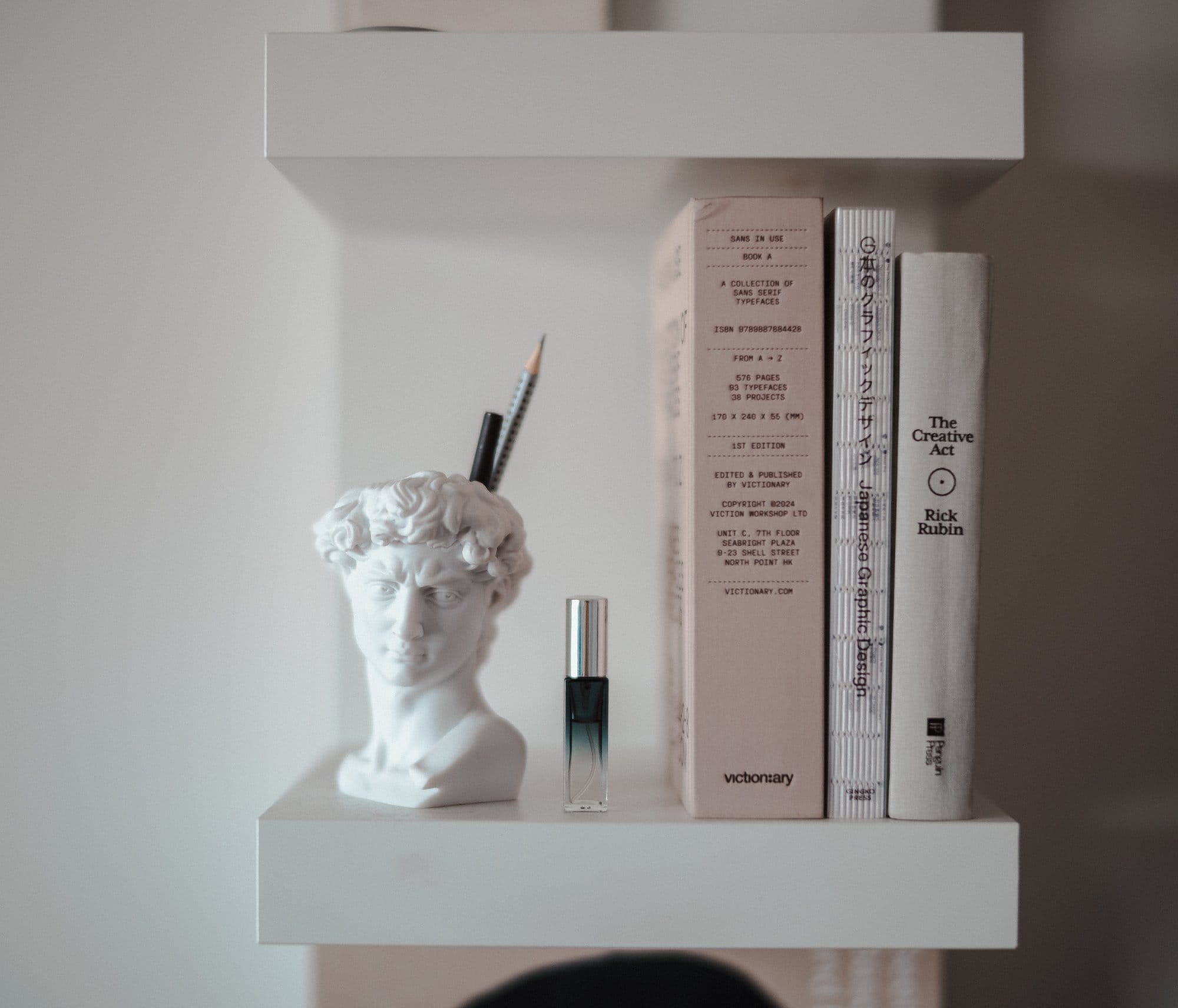 A minimalist white shelf holding a head-shaped pencil holder, a small glass bottle, and three books including The Creative Act by Rick Rubin and Sans in Use by Victionary