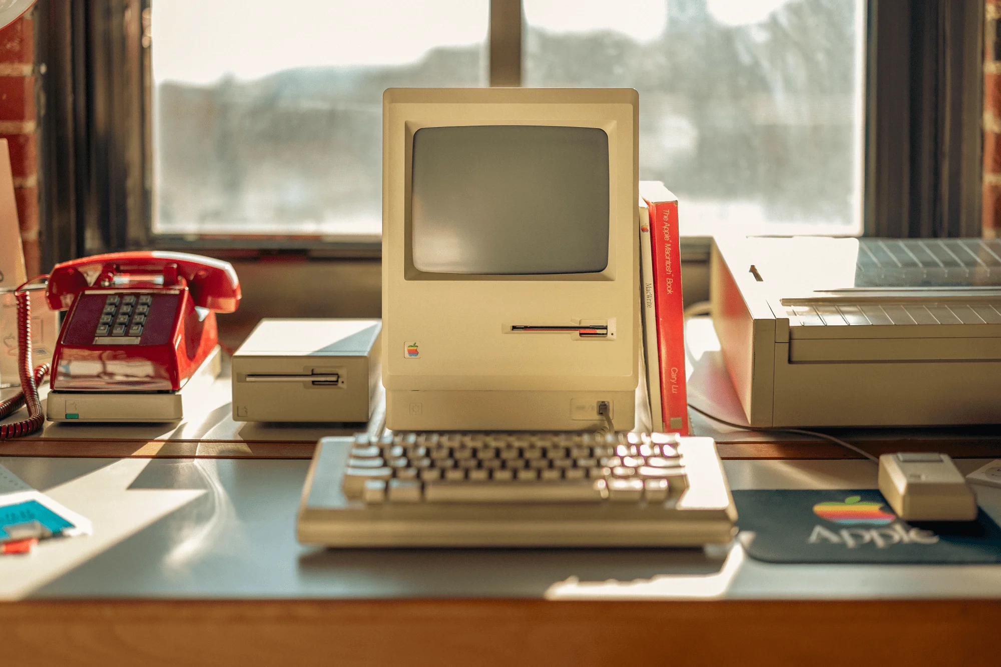 A vintage Apple desk setup with an all-in-one Macintosh (M0001), a Macintosh keyboard (M0110), a Macintosh mouse (M0100), a 400K external drive, an ImageWriter printer, and a red rotary phone, with The Apple Macintosh Book and MacWrite manual next to the monitor