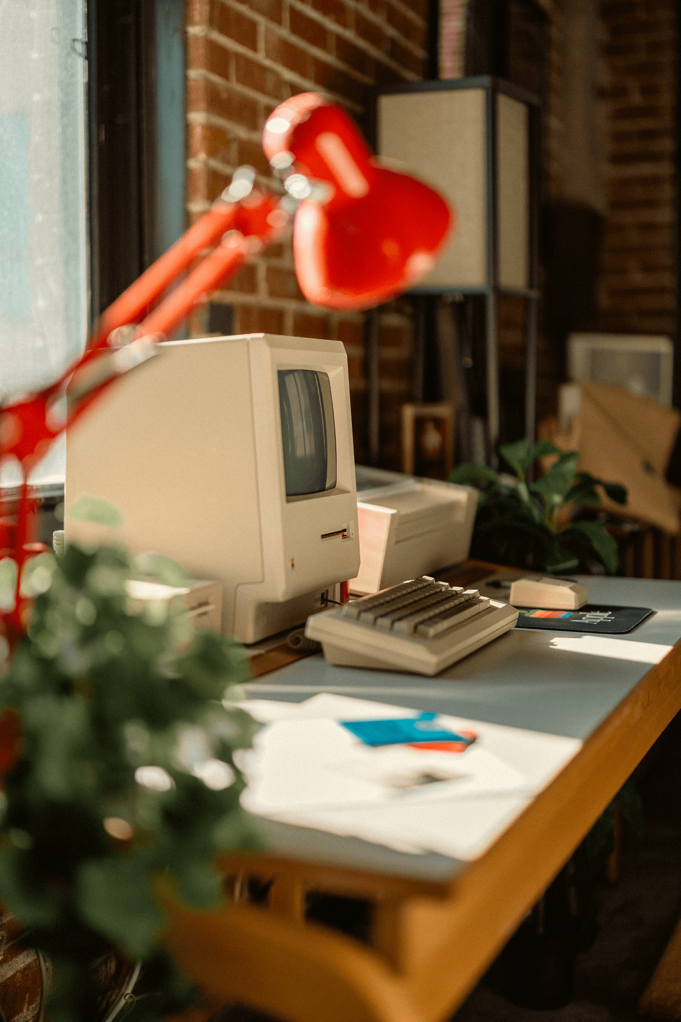A side view of a vintage Apple desk setup with an all-in-one Macintosh (M0001), a Macintosh keyboard (M0110), a Macintosh mouse (M0100) on an Apple mouse mat, a 400K external drive, and an ImageWriter printer, with a red desk lamp and green plants nearby