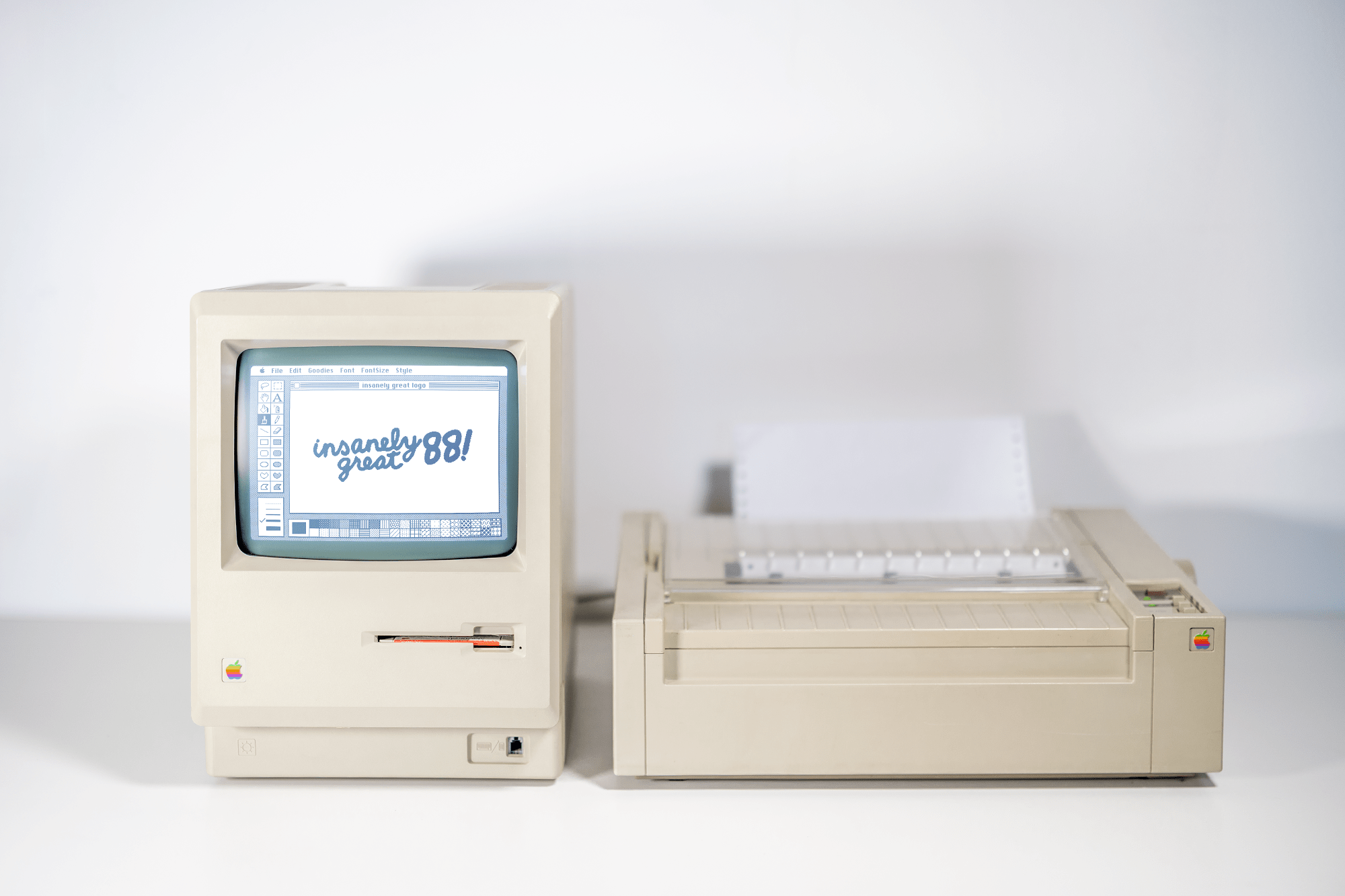 A front-facing view of an all-in-one Macintosh (M0001) displaying “insanely great 88!” next to an ImageWriter printer against a plain white background
