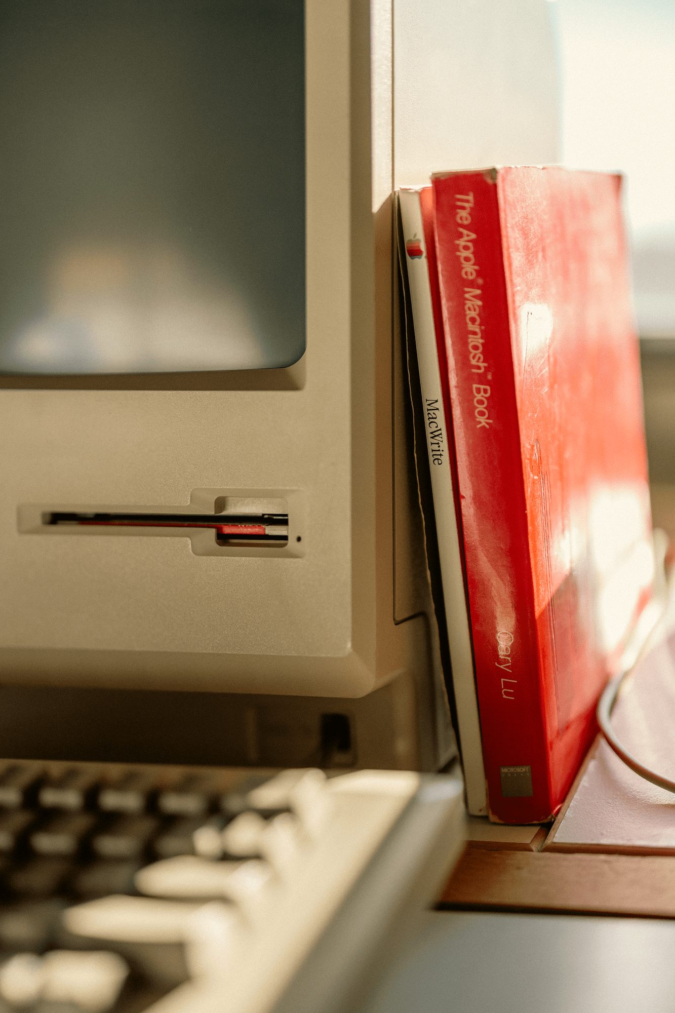 A close-up of an all-in-one Macintosh (M0001) with a floppy disk inserted, next to two books leaning against its side — The Apple Macintosh Book by Cary Lu and a MacWrite manual