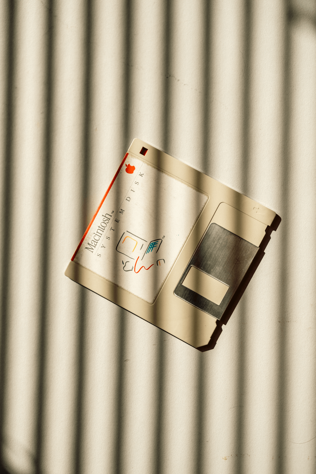 A Macintosh system disk lying on a light-coloured surface with shadow stripes falling across it