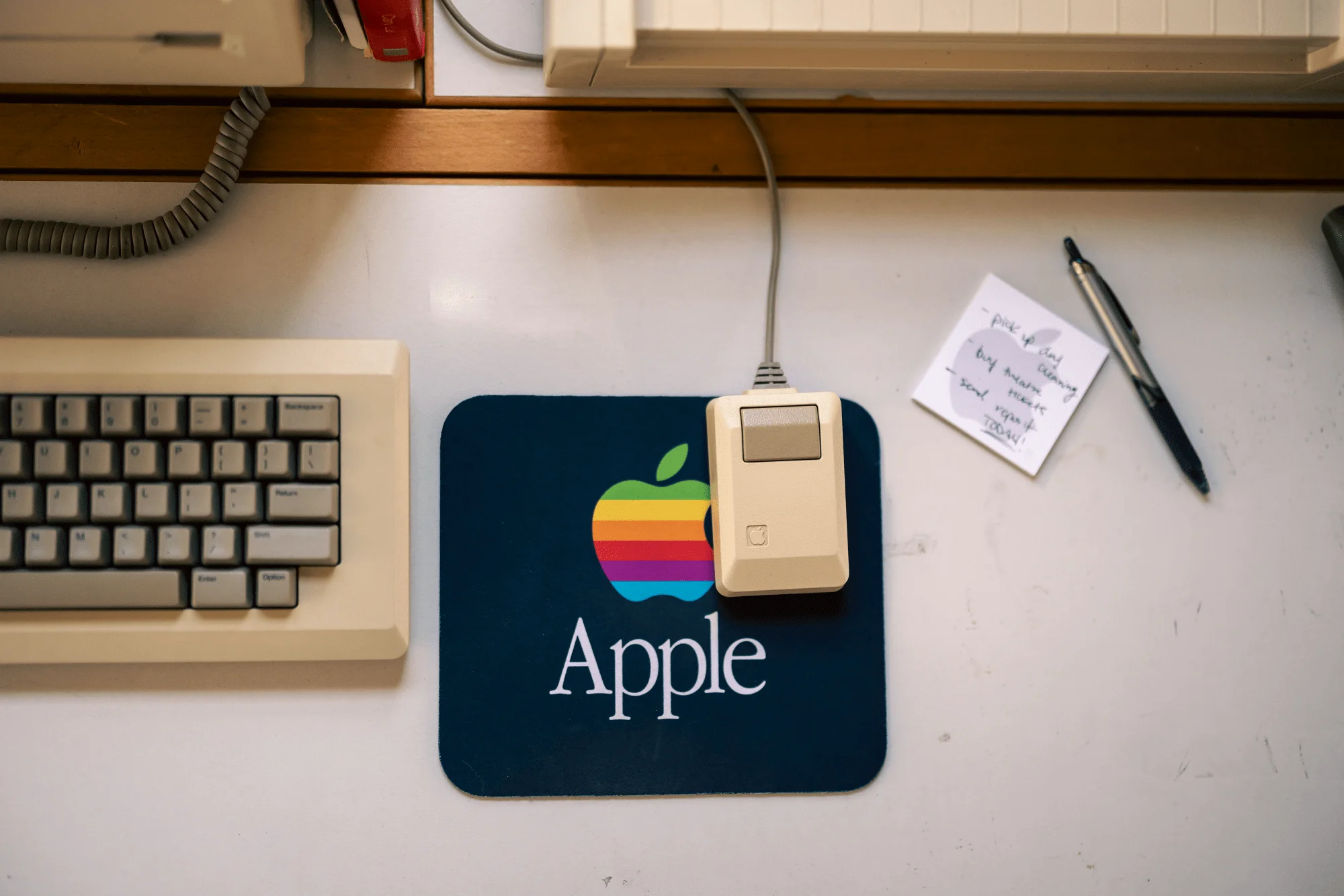 A close-up of a vintage Apple desk setup with a Macintosh mouse (M0100) on a rainbow Apple mouse mat, part of a Macintosh keyboard (M0110), a handwritten sticky note, and a pen on a white desk surface